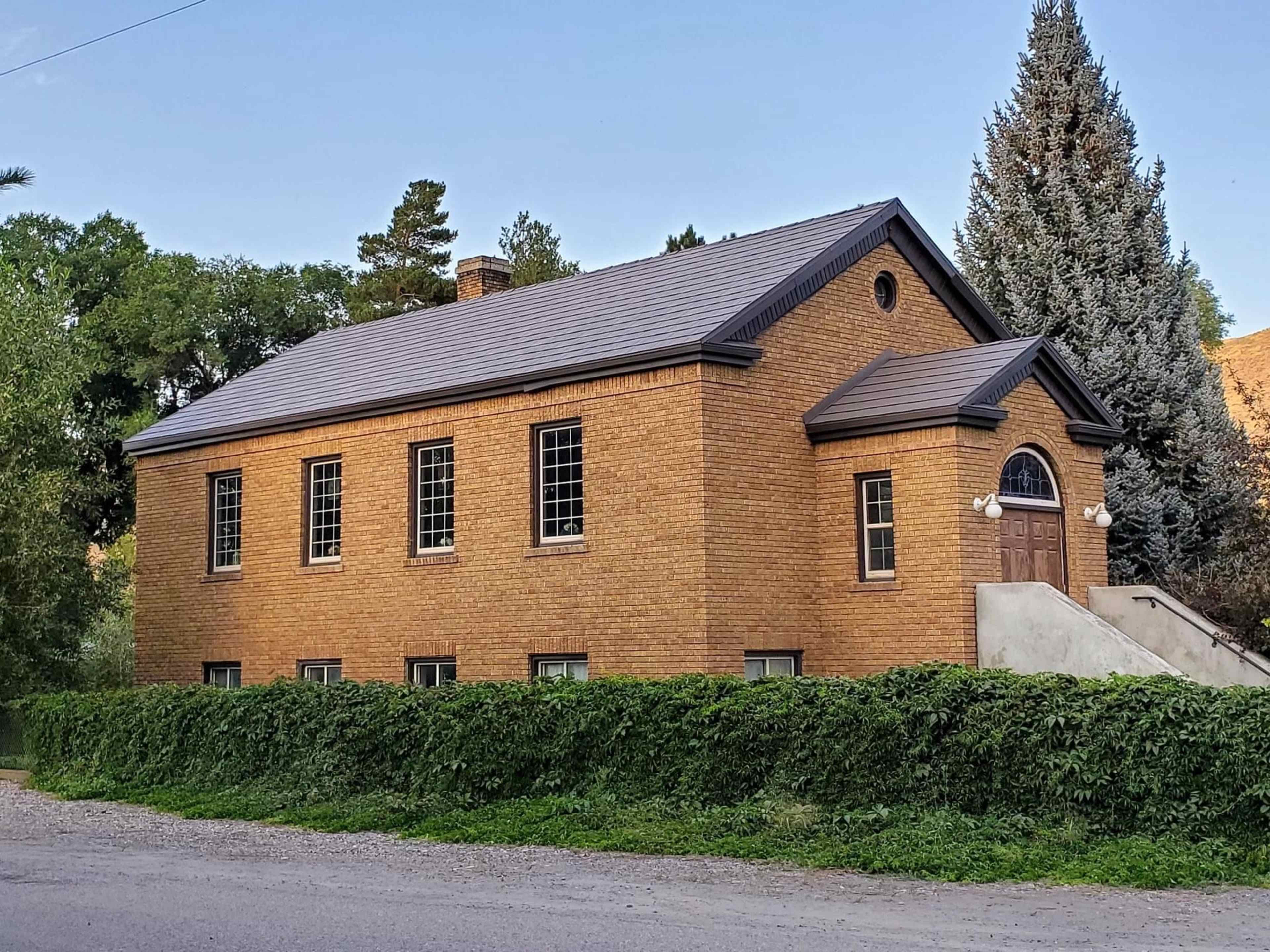A brick building with a sloped roof, featuring multiple windows and a set of steps leading to a front entrance, surrounded by a low hedge.