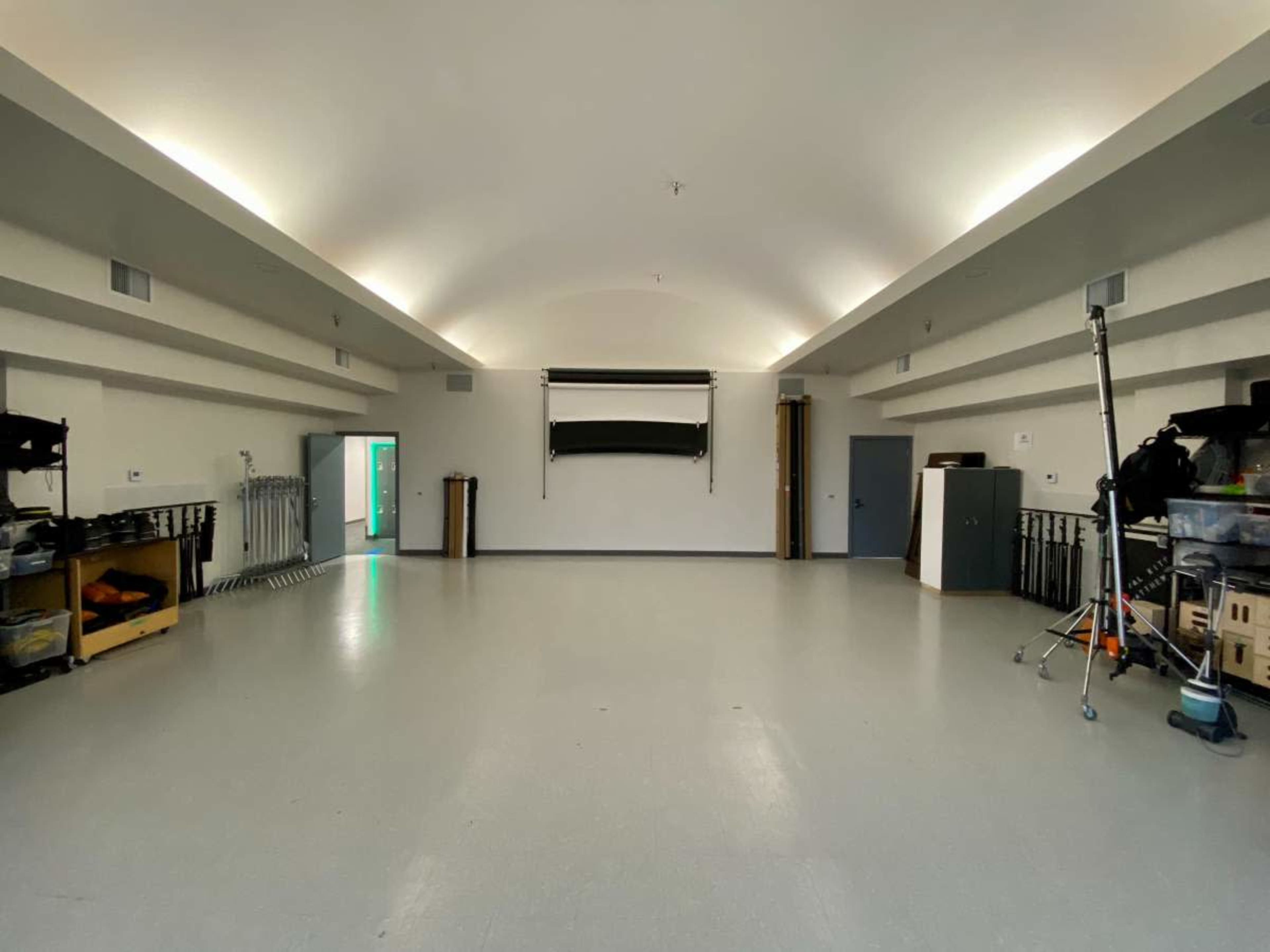 The image shows a spacious, empty room with high ceilings, a large white wall, and minimal furniture against the sides.