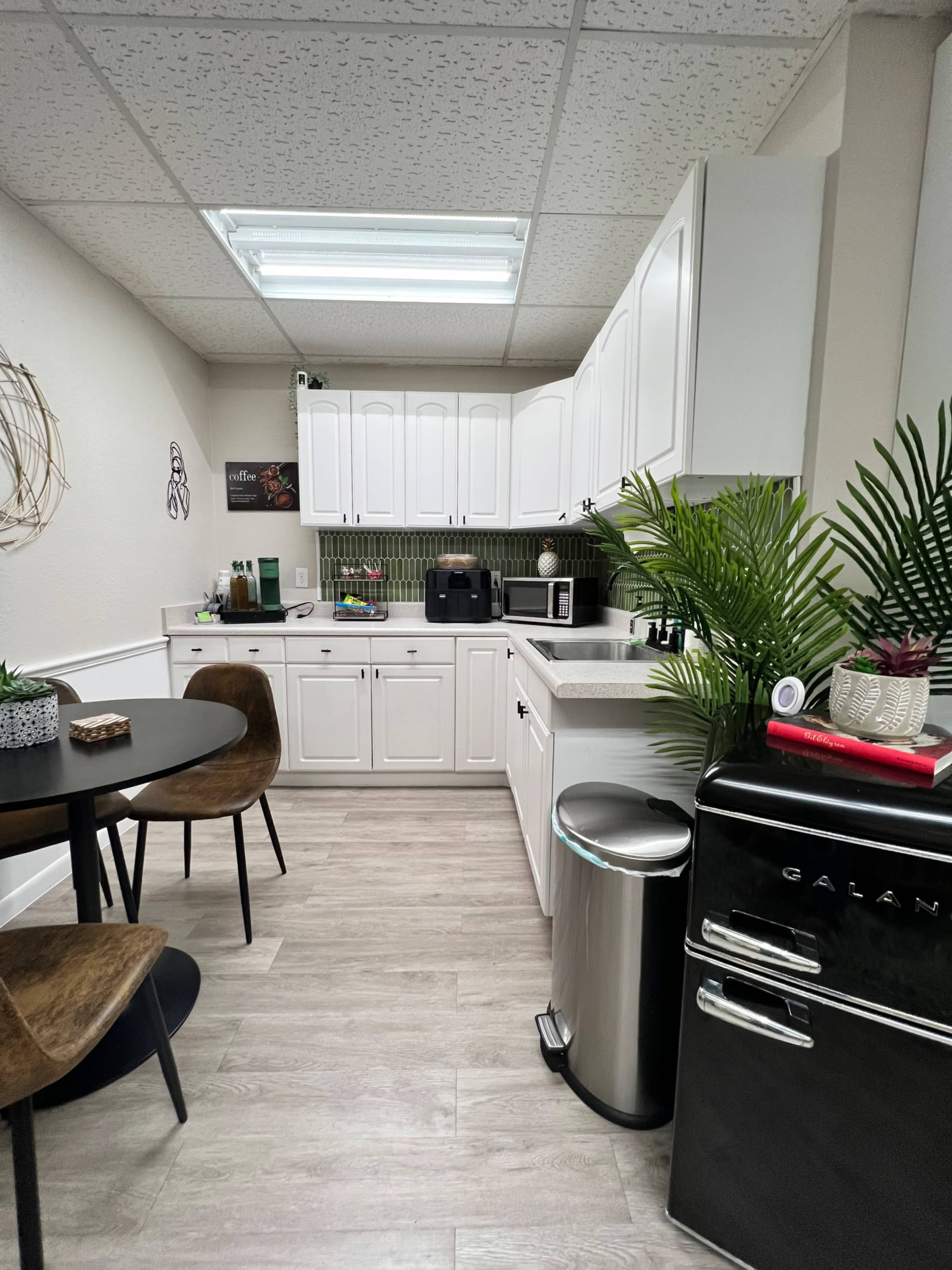 The image shows a modern kitchen with white cabinetry, a small round dining table, and appliances including a refrigerator and coffee maker.