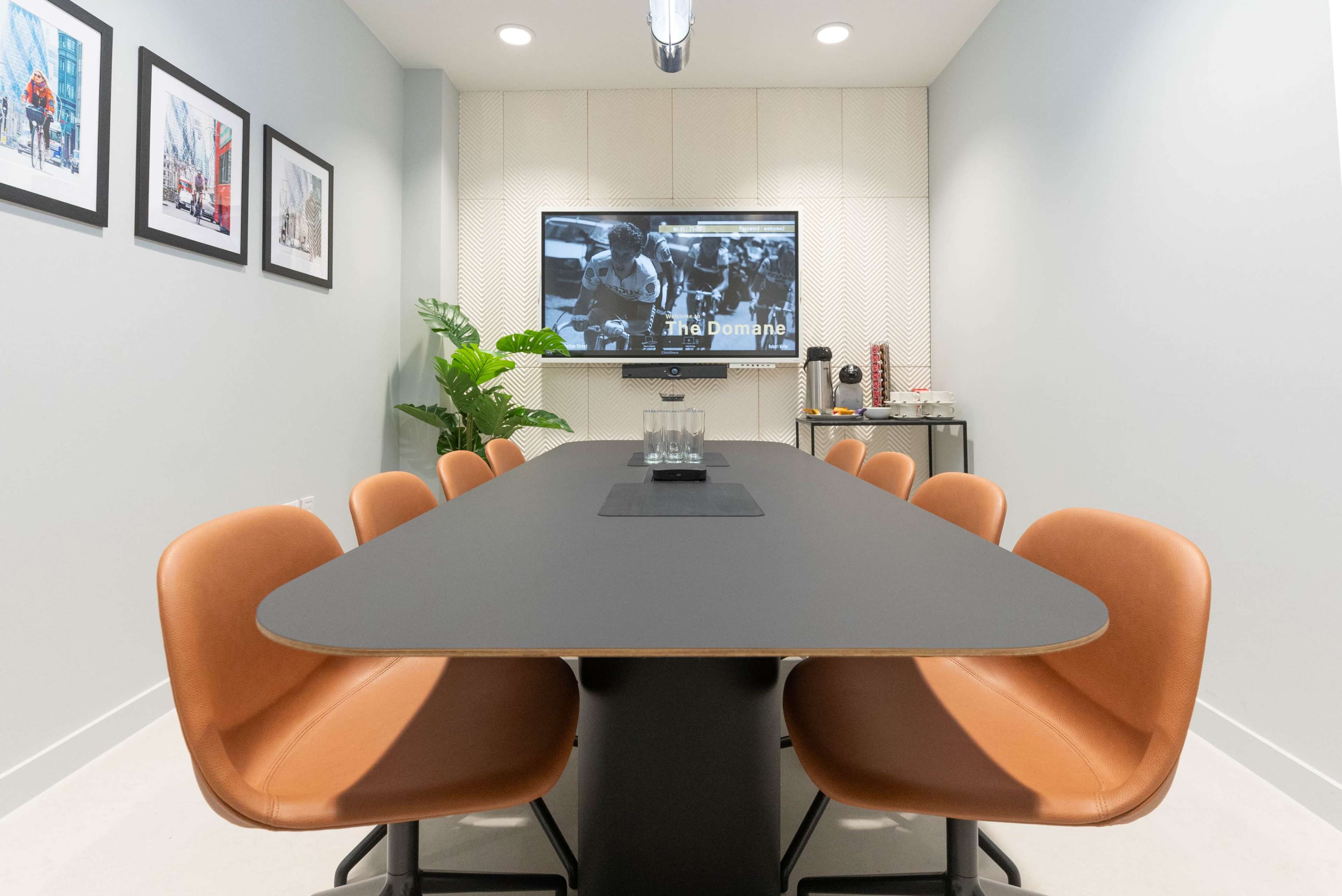 A modern conference room features a long black table surrounded by brown leather chairs, with a wall-mounted television displaying an image and framed photographs in the background.