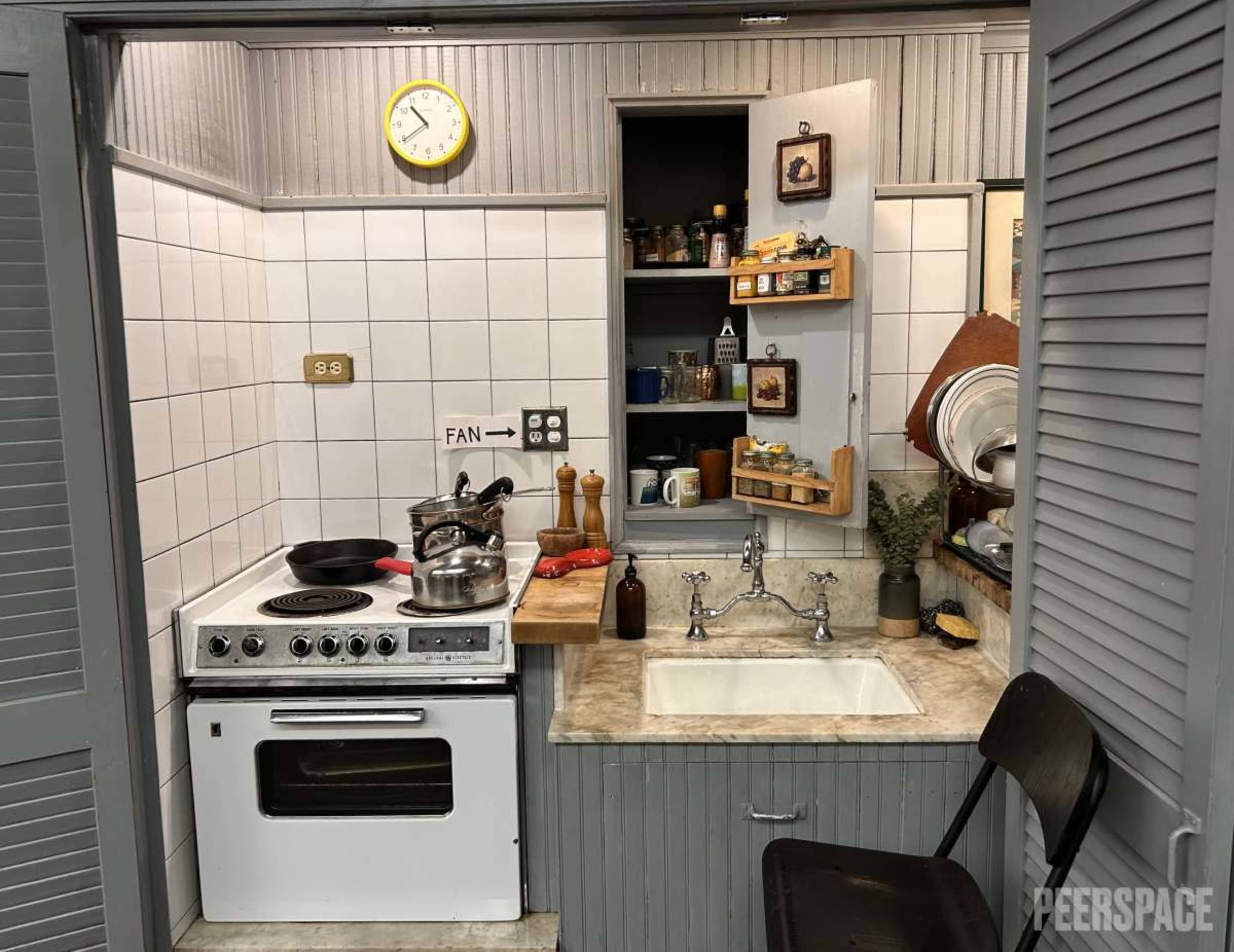 The image shows a compact kitchen with a stove, sink, and wooden shelves lined with various kitchen items.