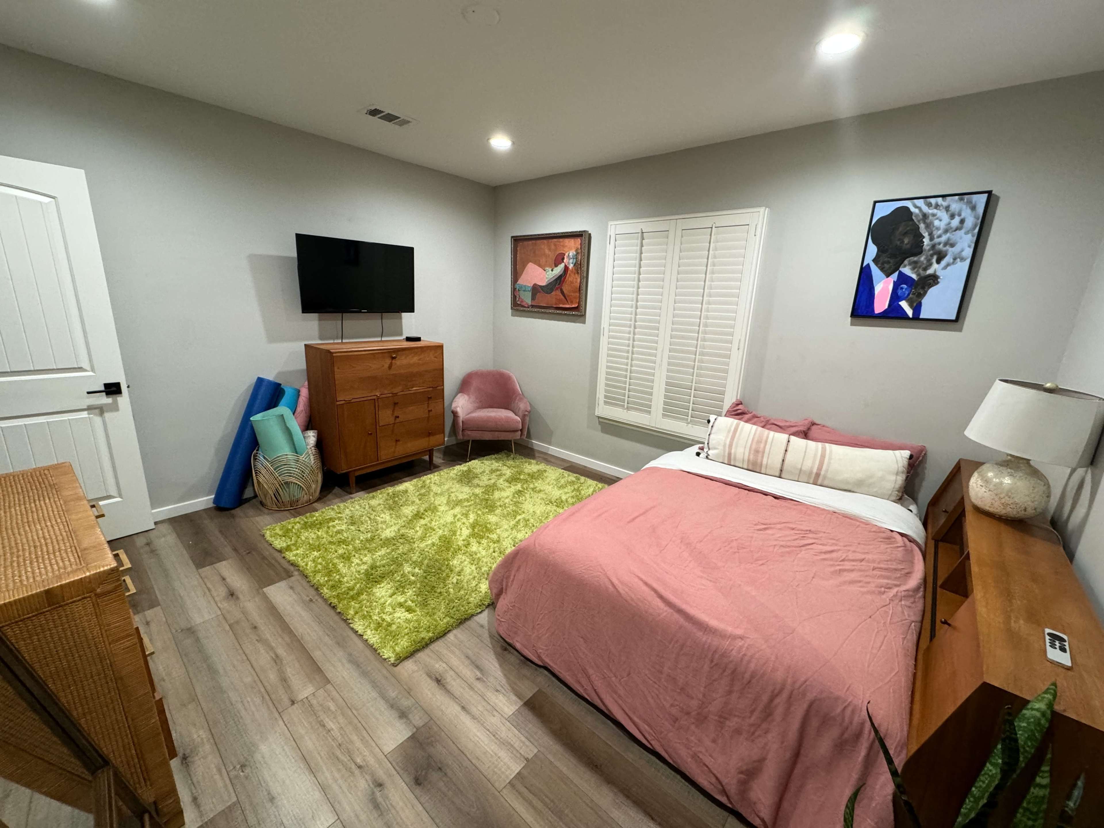The image shows a neatly arranged bedroom featuring a pink bedspread, a wooden dresser, a television mounted on the wall, and a light green rug on the floor.