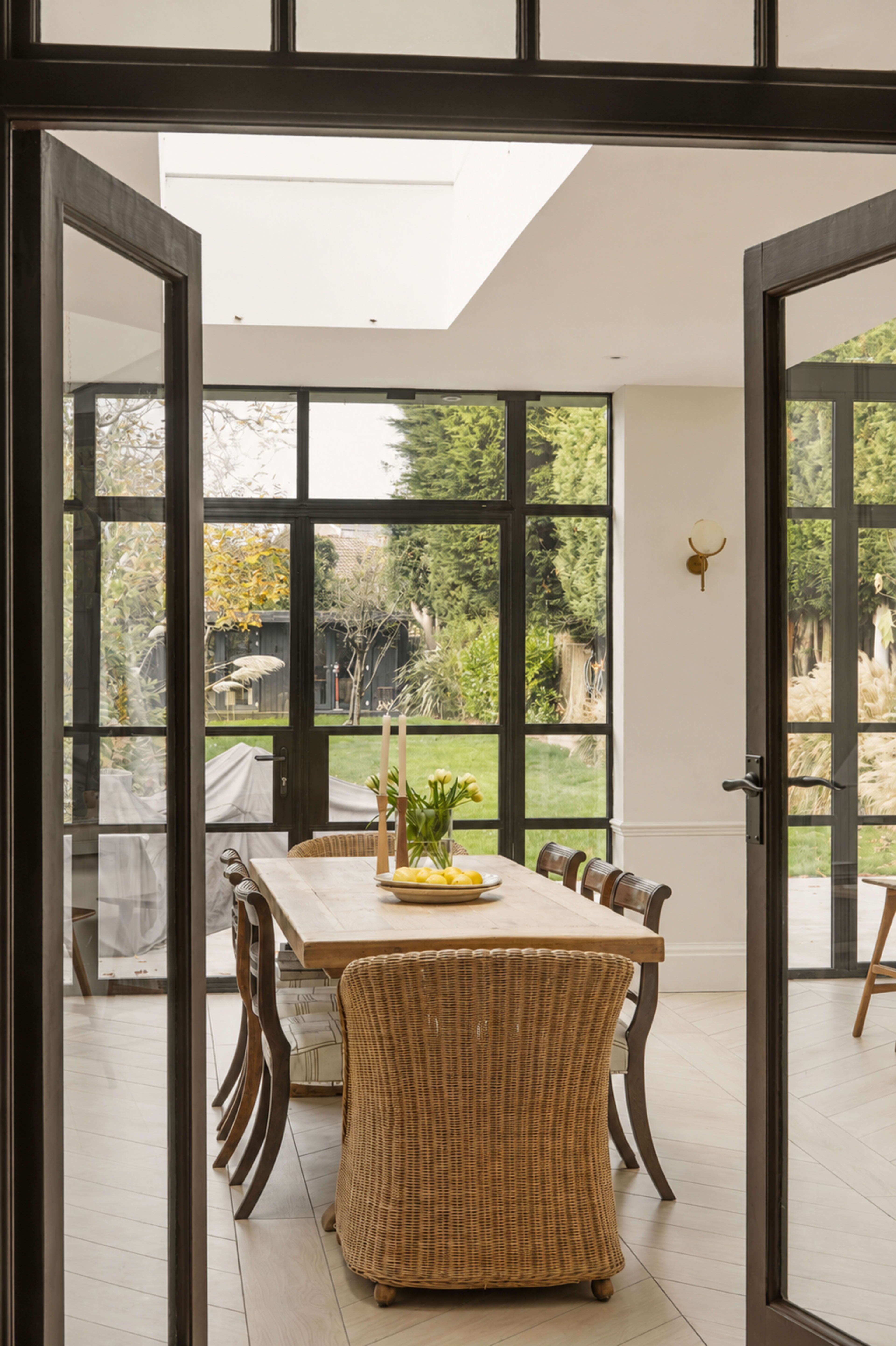 A dining area features a wooden table surrounded by chairs, with large glass doors leading to a garden outside.