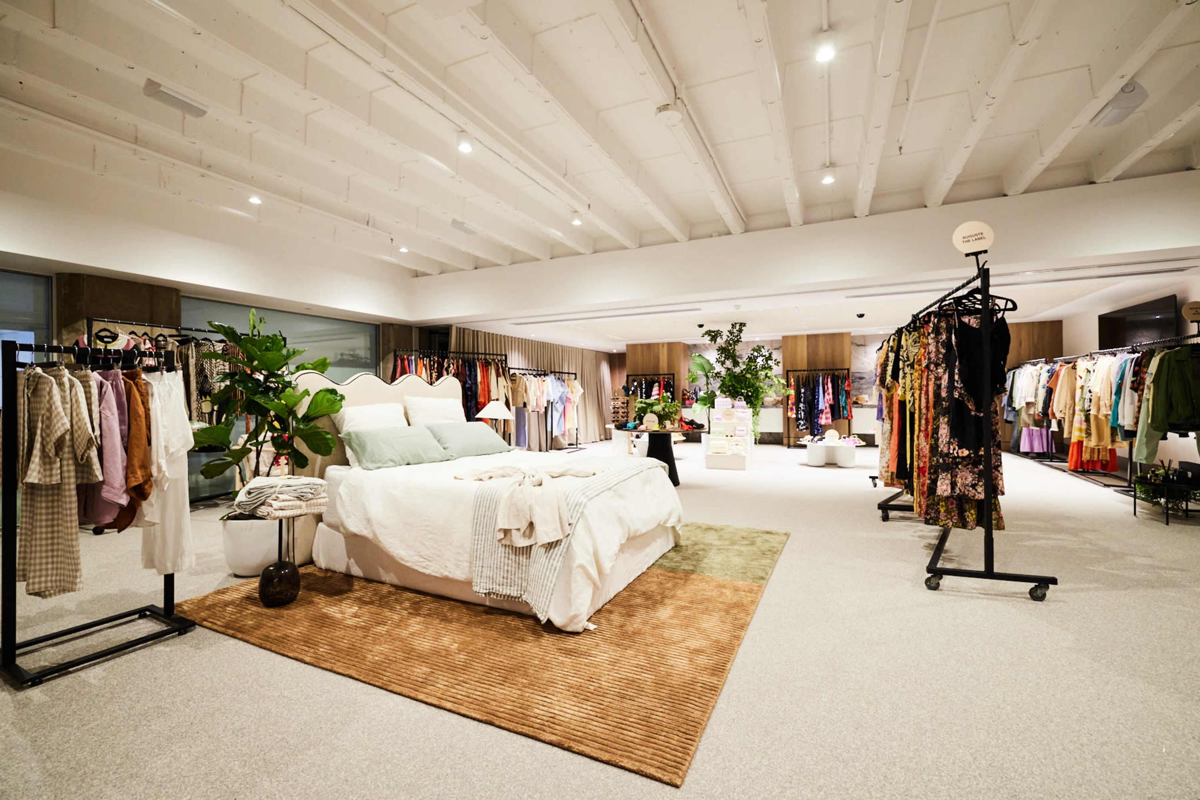 A spacious clothing store featuring a well-decorated display area with a bed, along with multiple racks of clothing and potted plants.