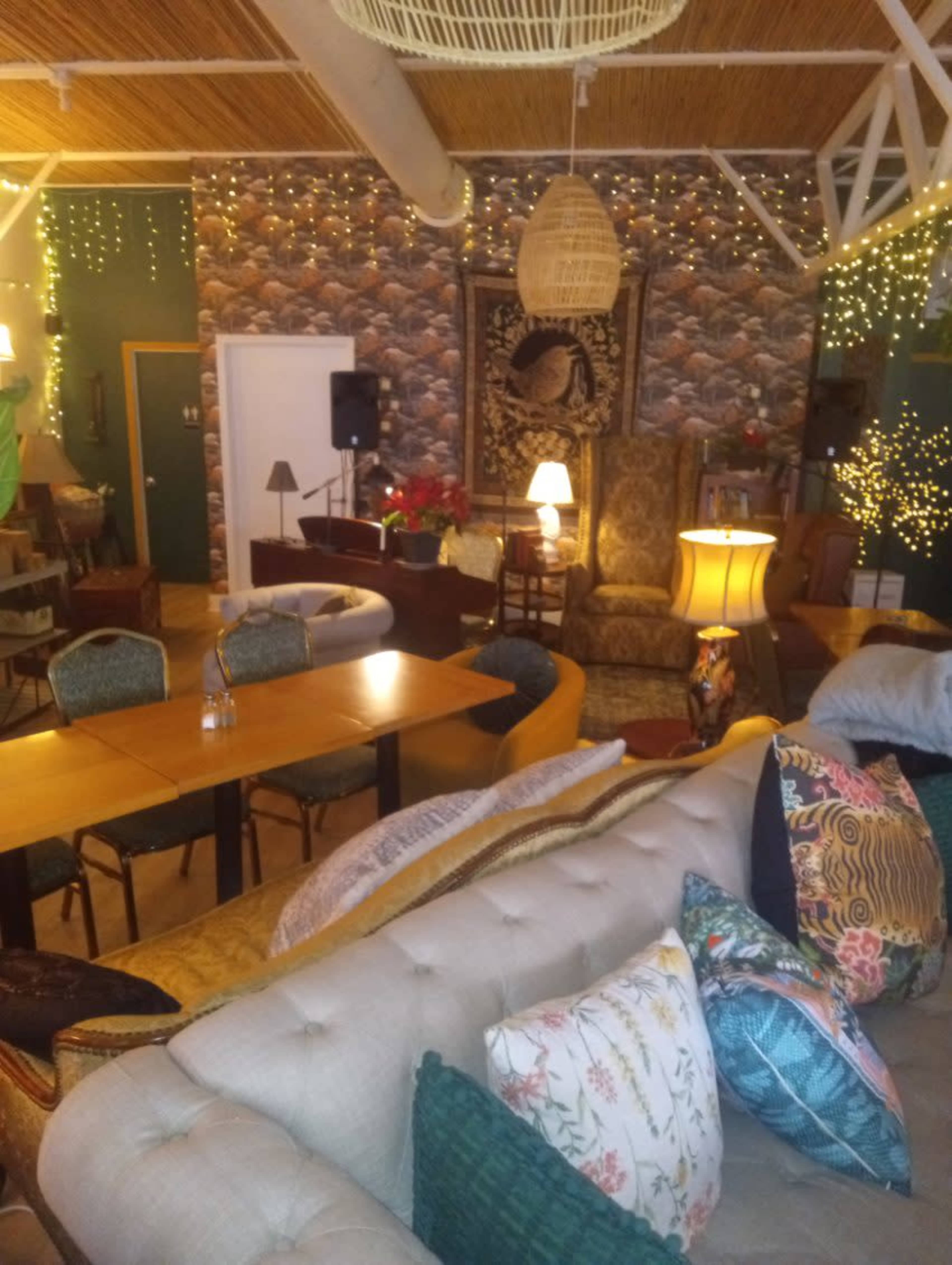 The image shows a cozy interior space filled with various seating arrangements, warm lighting, and decorative elements like plants and artwork.