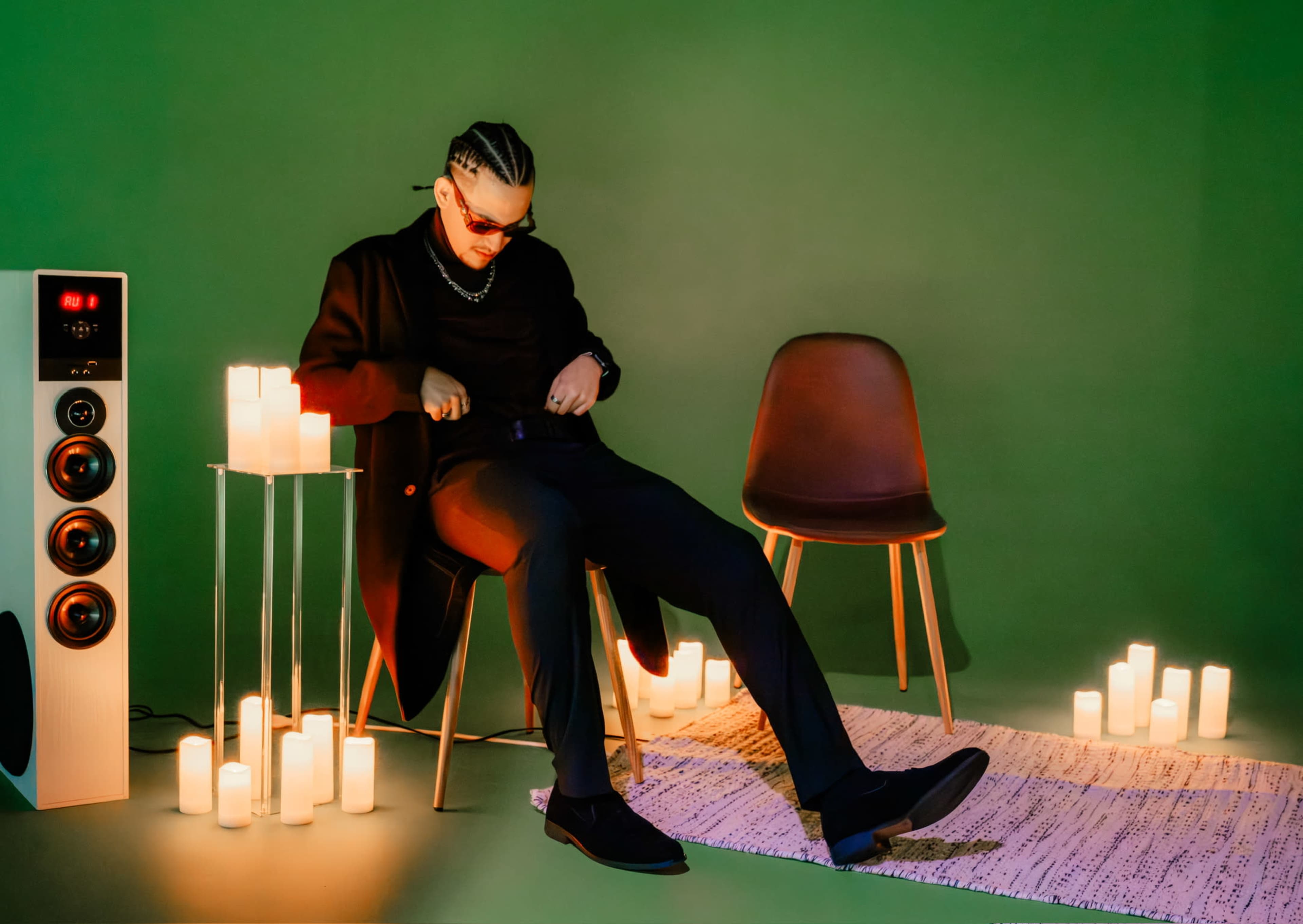 A person sits on a chair in a dimly lit room surrounded by candles and a speaker, adjusting their clothing.