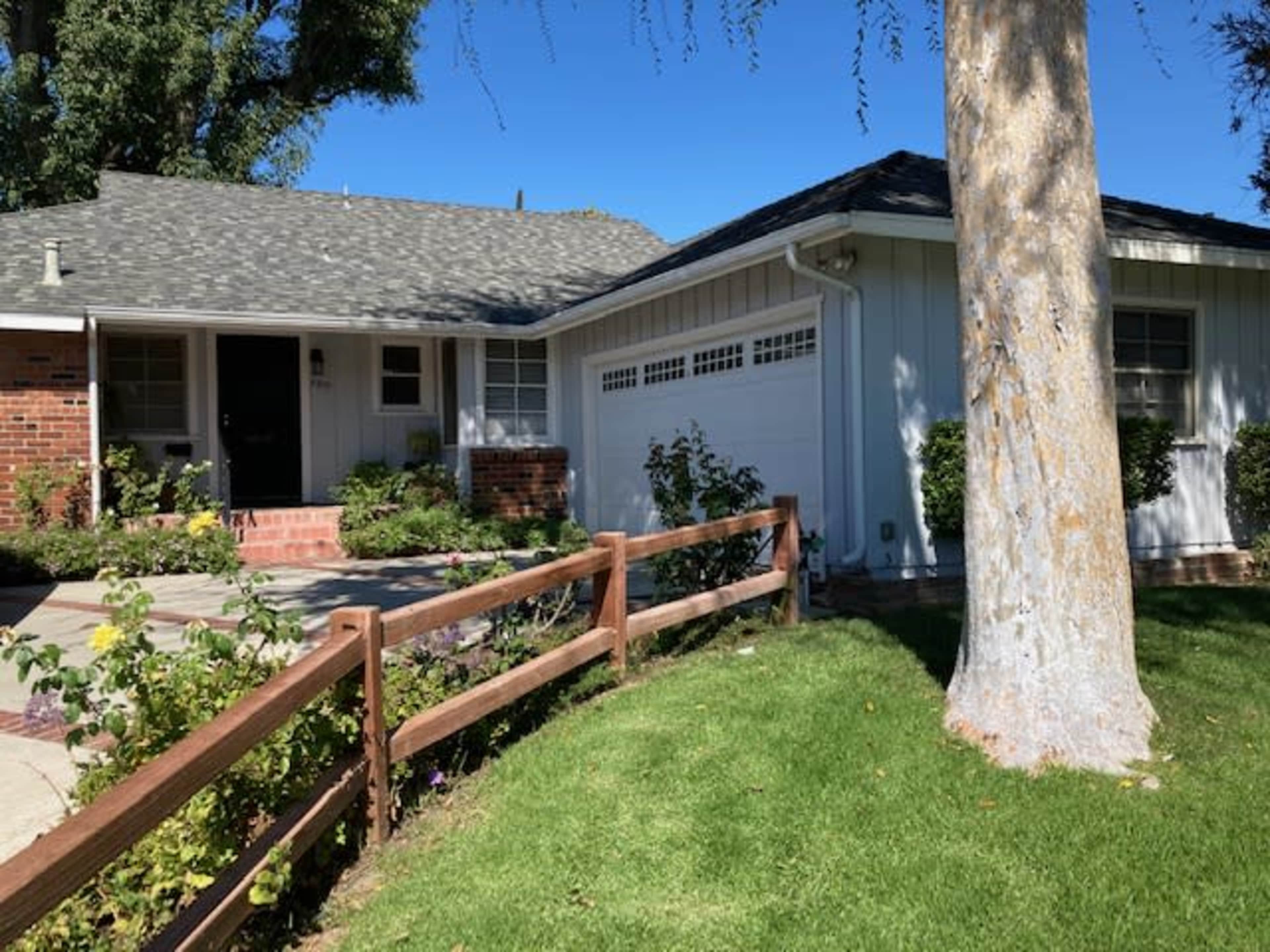 A single-story house with a shingled roof and a white exterior is surrounded by a manicured lawn and a wooden fence.