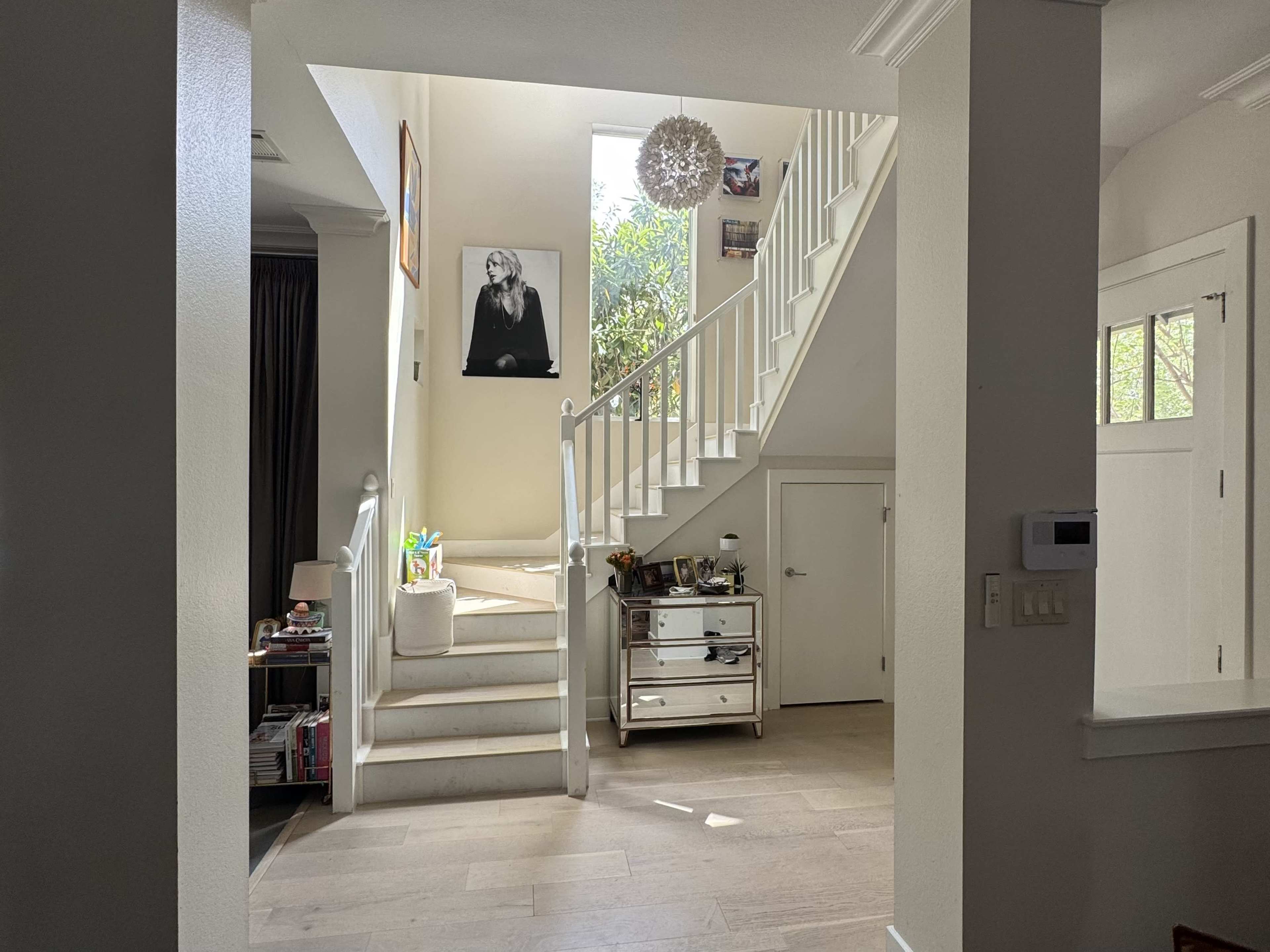 A bright entryway features a staircase with a large window above, flanked by plants and framed artwork on the walls.