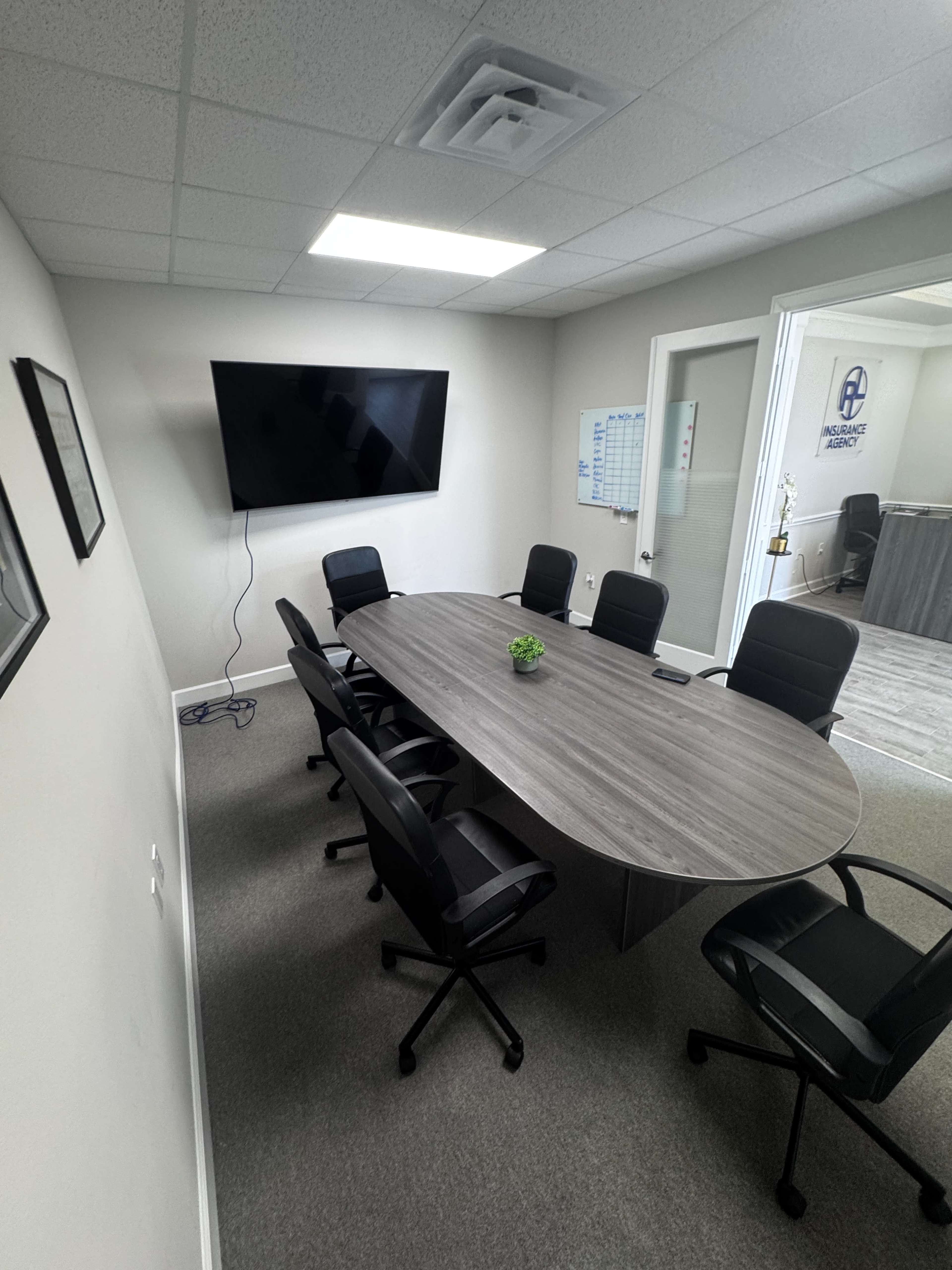 A modern conference room features a long oval table surrounded by black office chairs and a wall-mounted television.