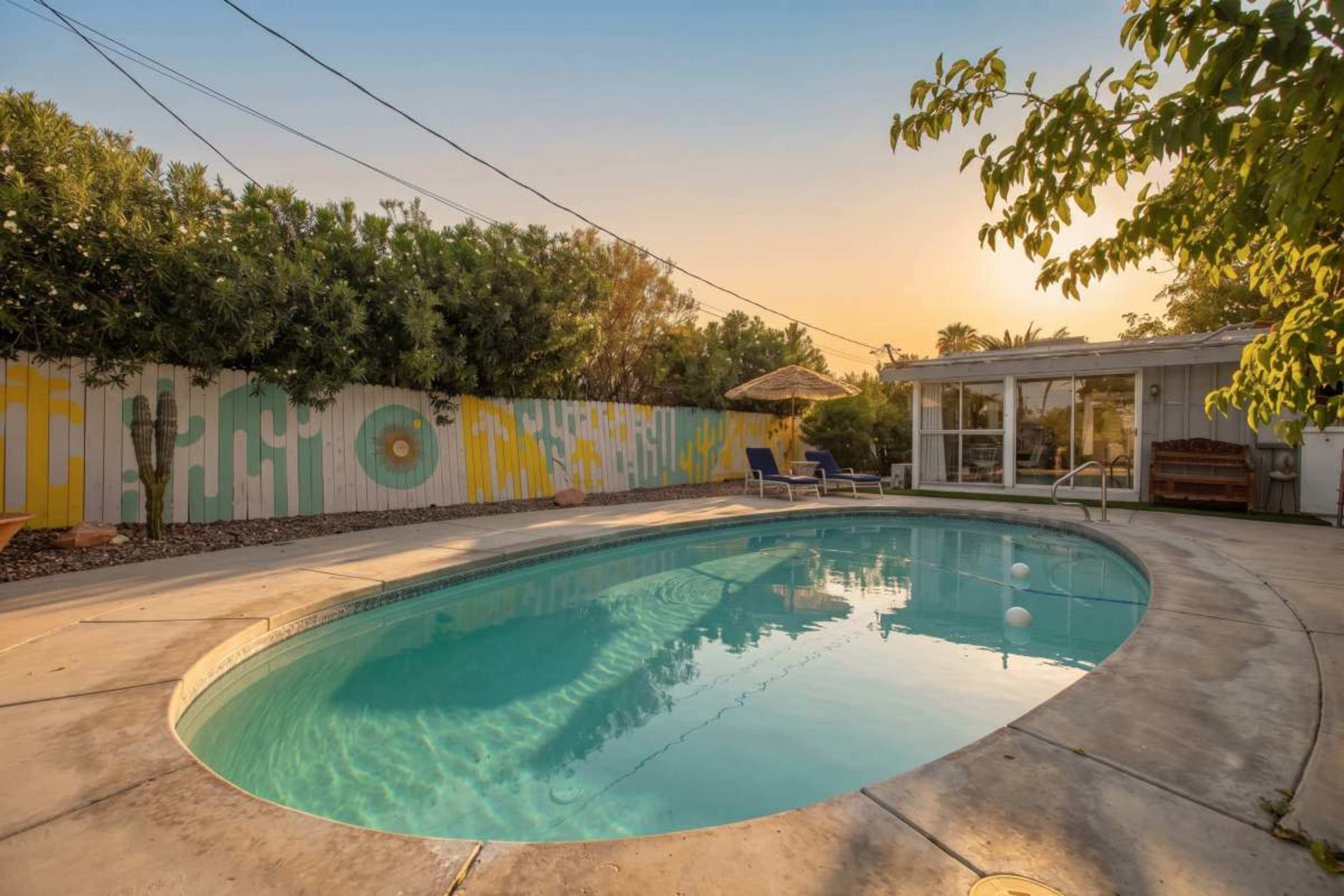 The image shows a swimming pool surrounded by a concrete deck and lush greenery, with a colorful mural on the fence in the background.