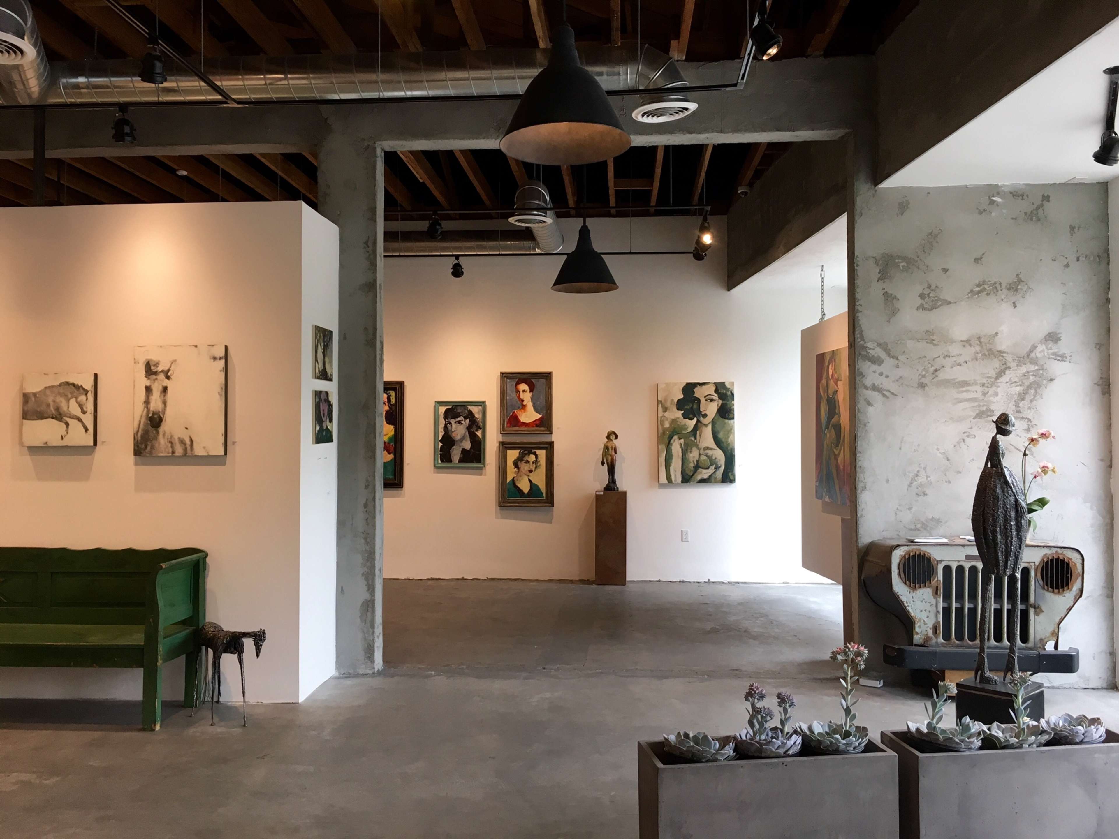 A modern art gallery featuring various paintings on the walls, a green bench, and sculptural displays in a spacious, well-lit environment with exposed ceilings and concrete flooring.