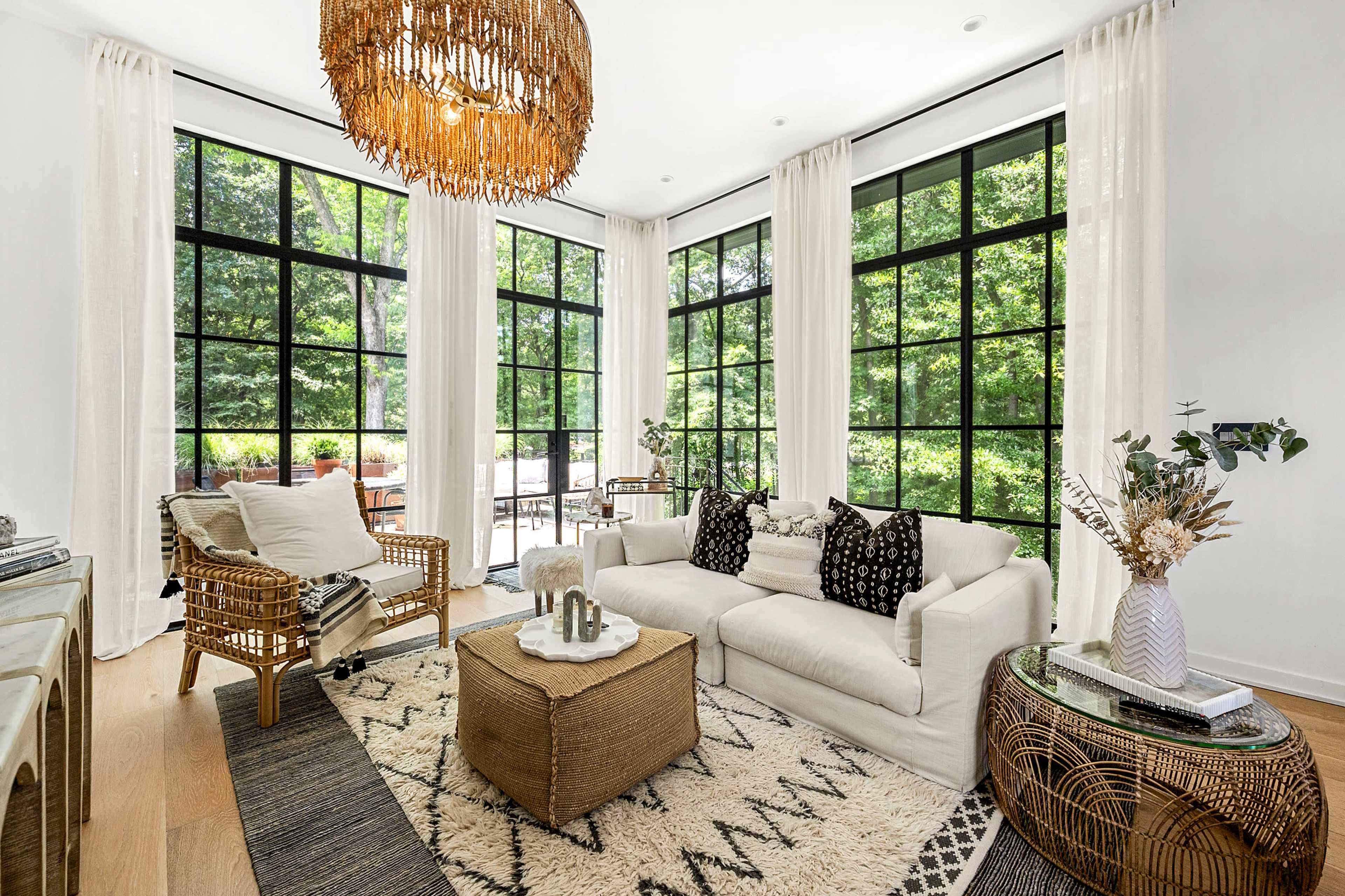 The living room features large windows that provide a view of a green exterior, with a white couch, a wicker coffee table, and decorative plants arranged in a warm, inviting space.