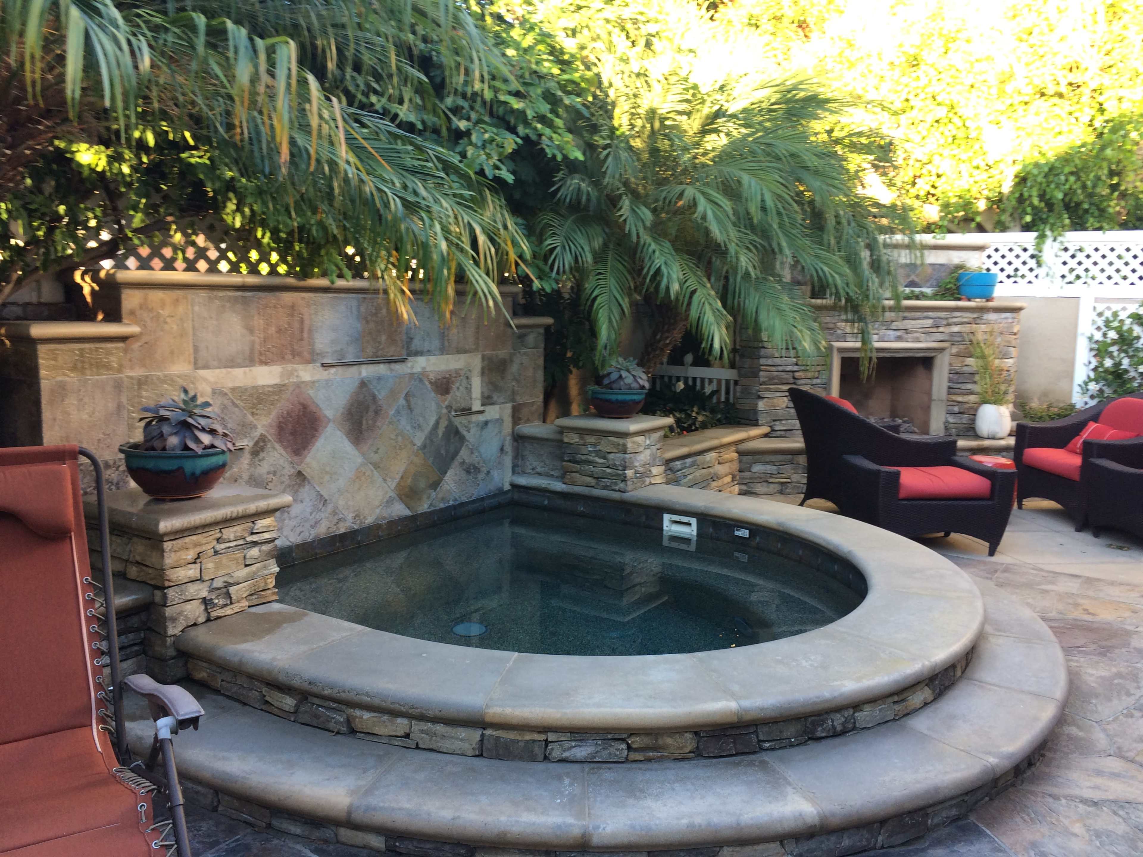 A circular hot tub surrounded by stone walls and tropical plants, with two black chairs and a terracotta-colored lounger nearby.