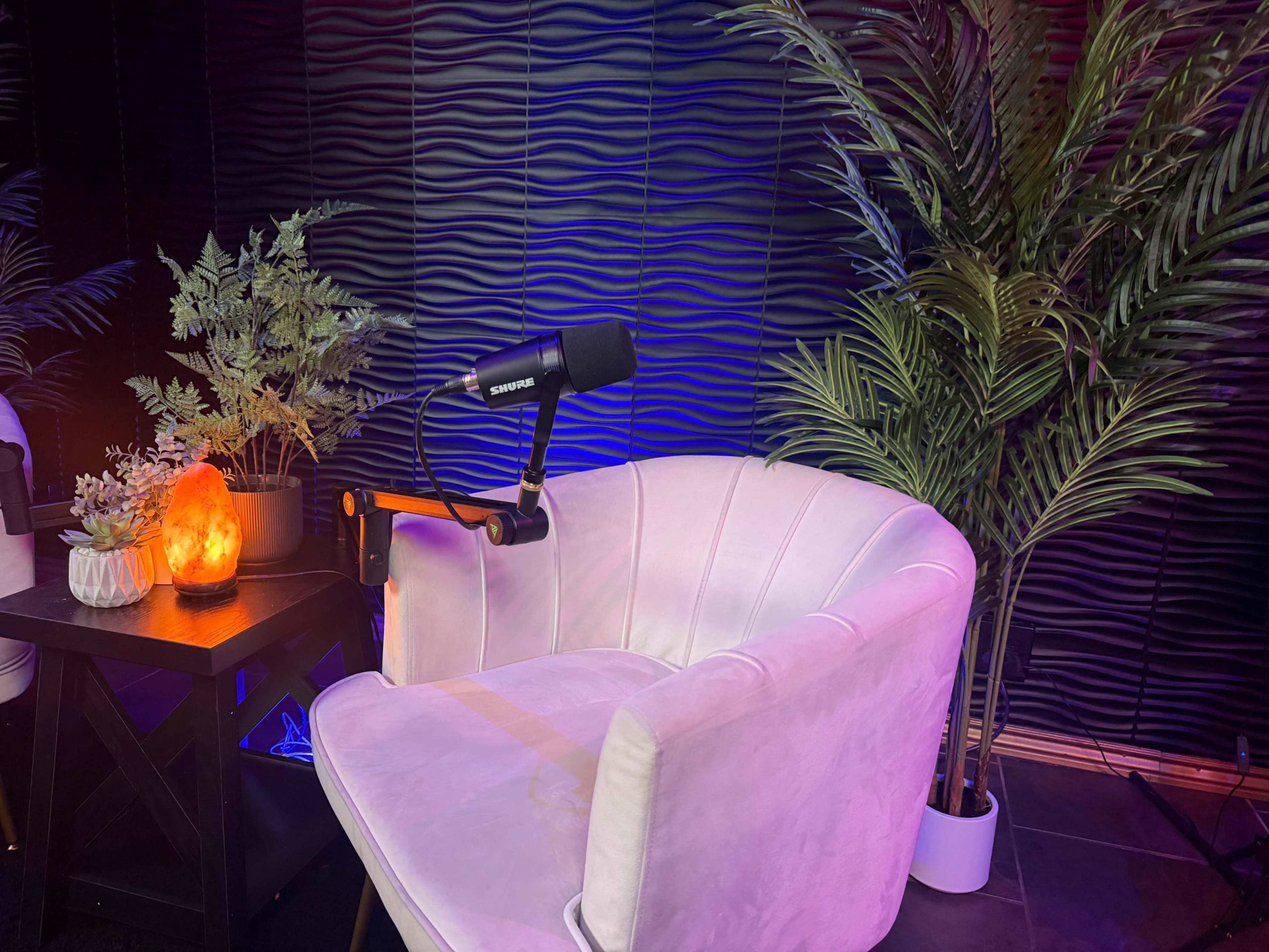 The image shows a microphone mounted on an arm positioned next to a white armchair, surrounded by decorative plants and soft lighting.