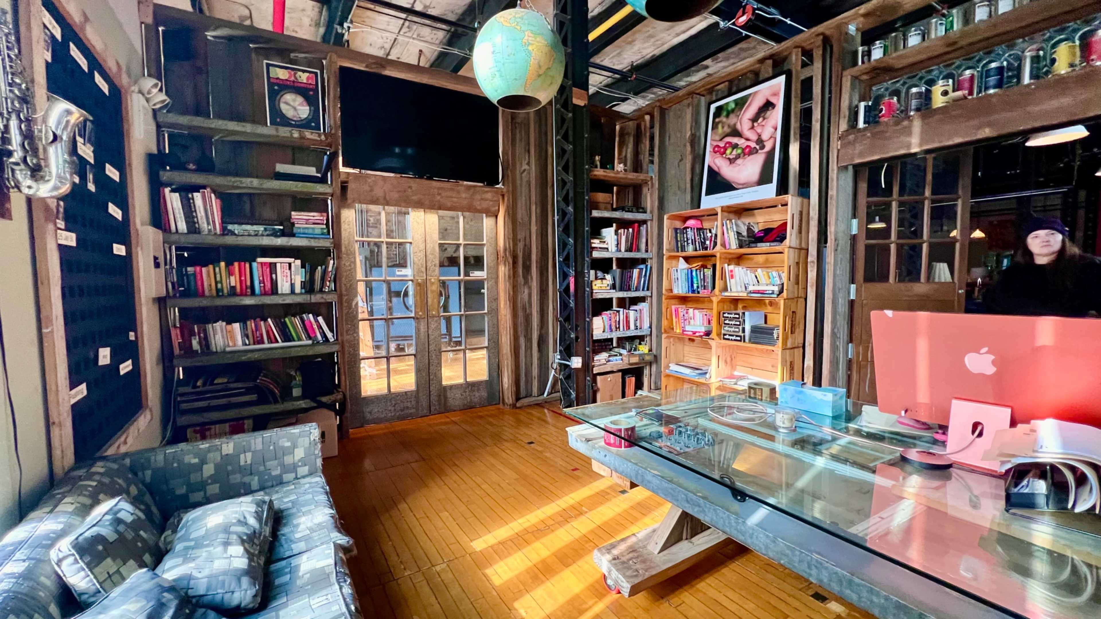 The image depicts a cozy, rustic interior of a library or co-working space, featuring wooden bookshelves filled with books, a glass table, and a couch, along with a large window.