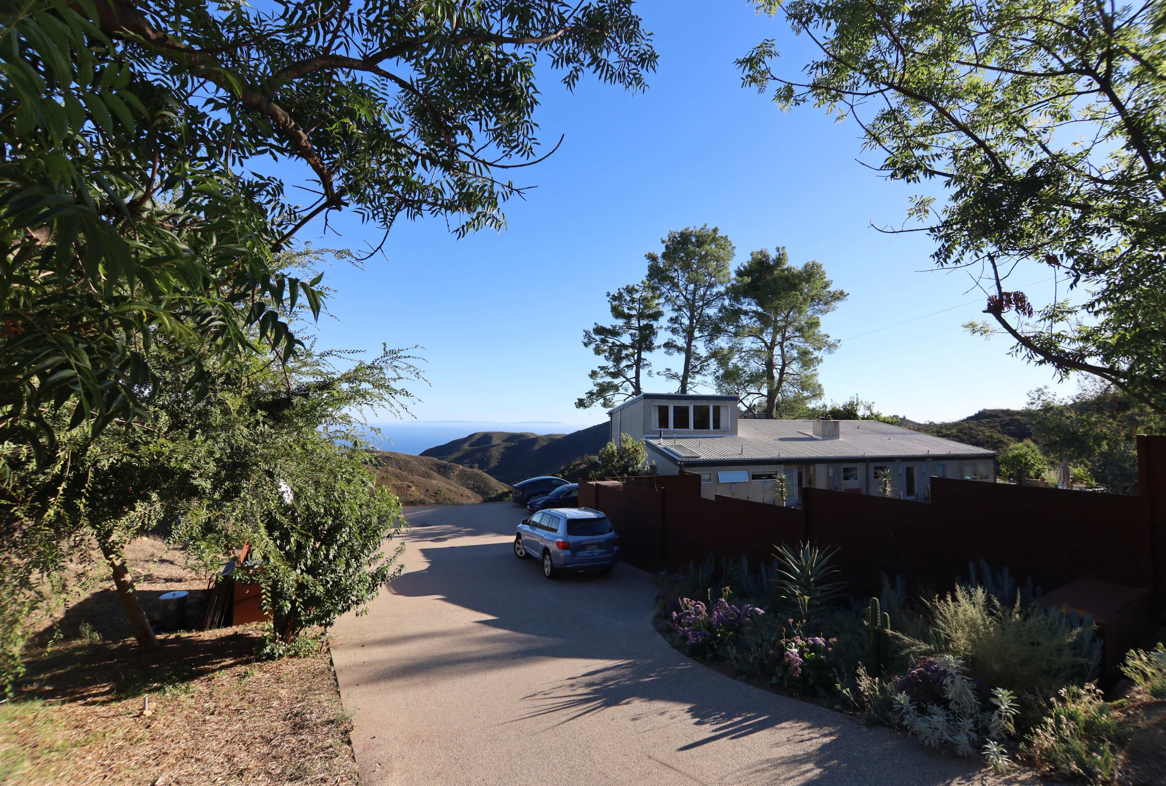 A house is situated on a hillside road overlooking the ocean, surrounded by trees and landscaped gardens.