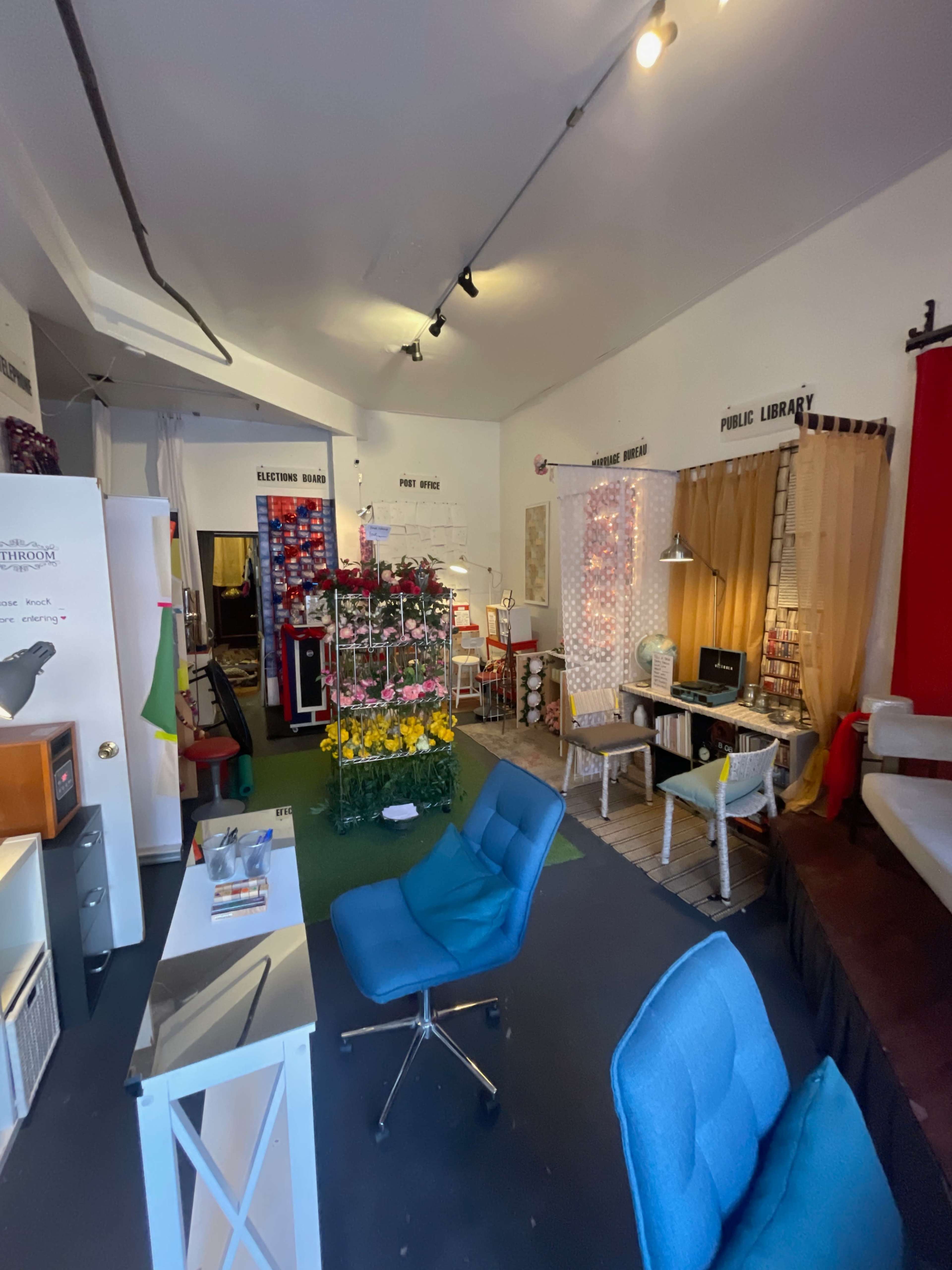 The image shows a creatively arranged room with various sections including a workspace, a wall of colorful items, and a seating area.