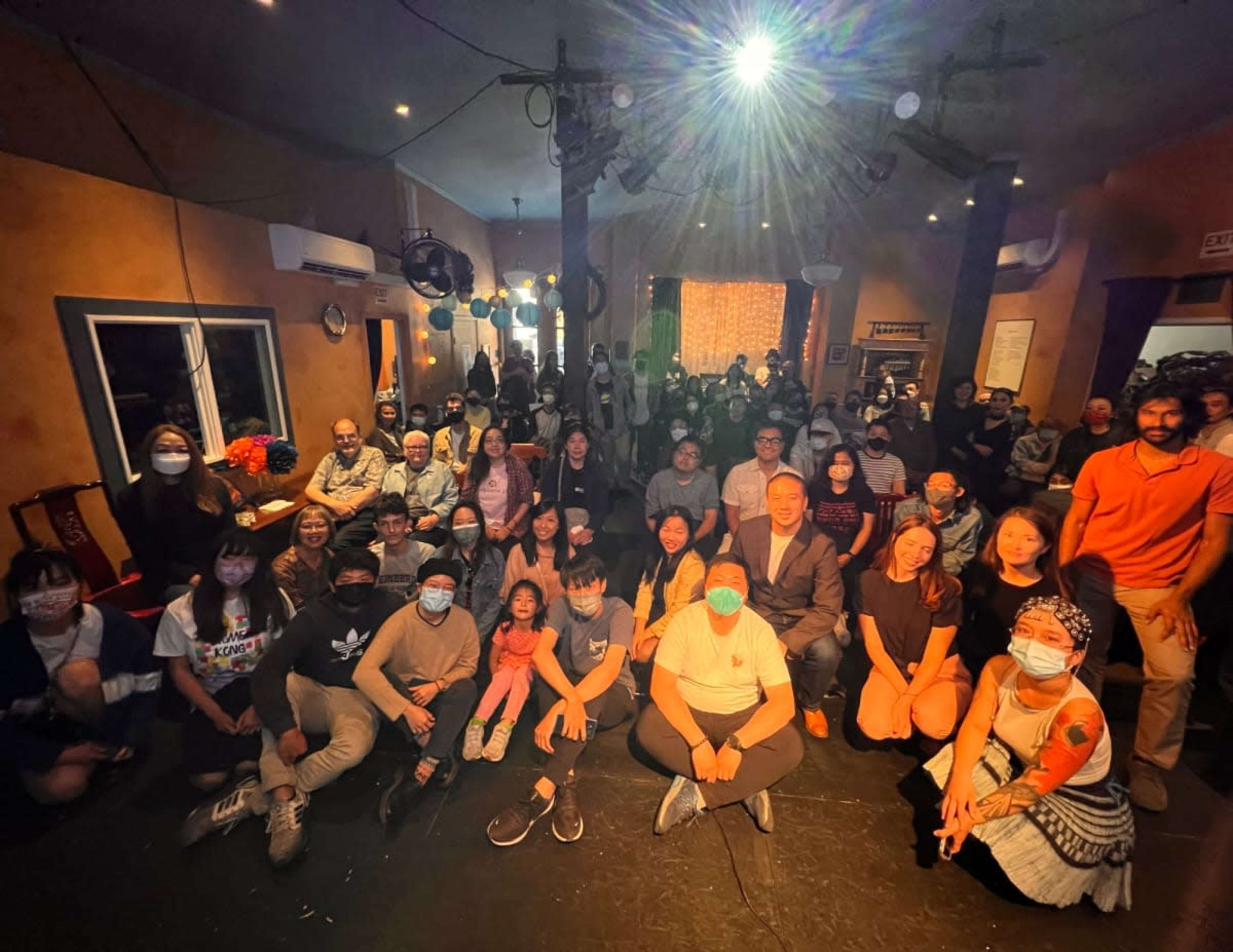 A large group of people poses together in a performance venue, with some individuals wearing masks and the audience visible in the background.