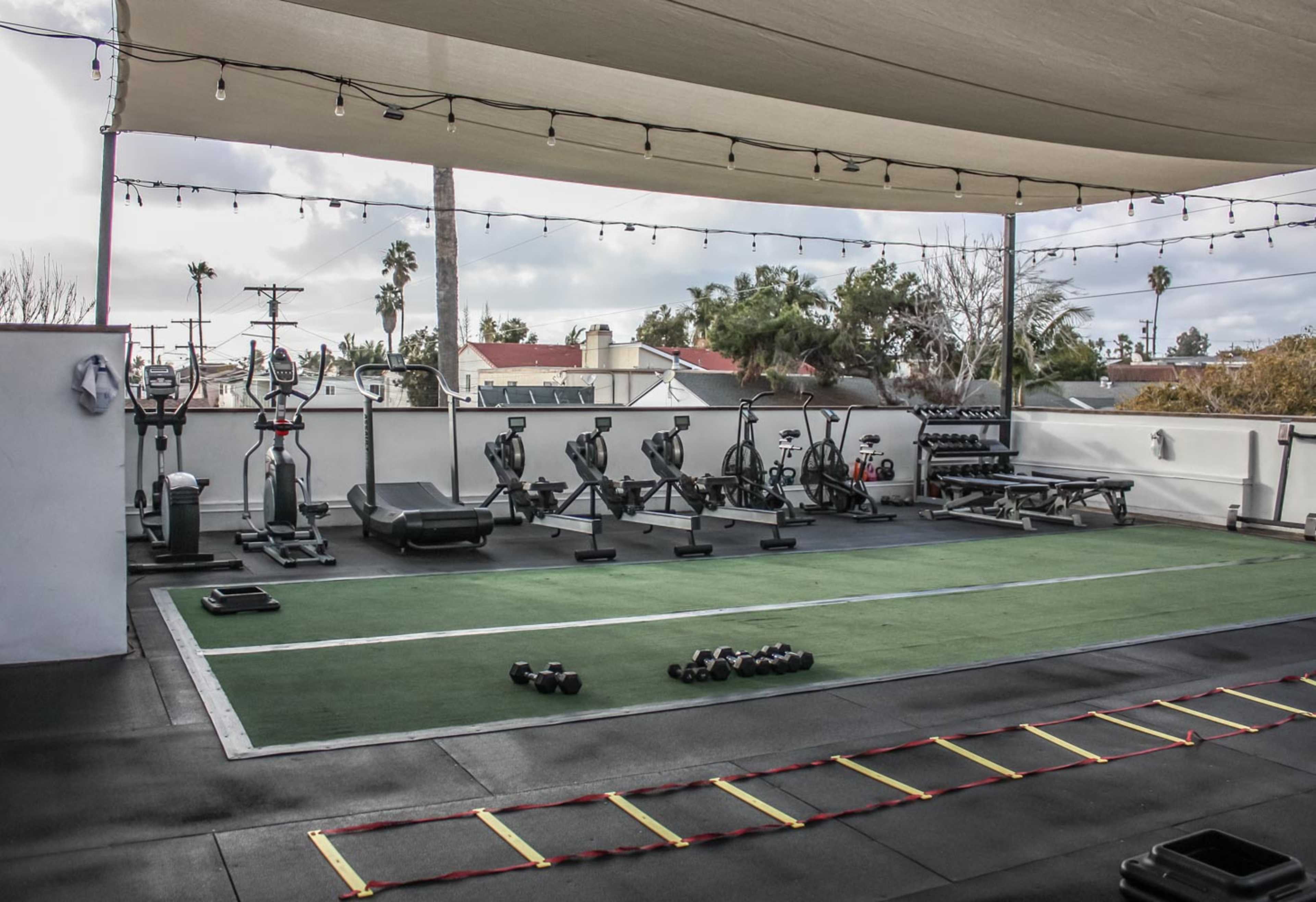 A rooftop gym equipped with various fitness machines, free weights, and exercise mats under a shaded canopy.