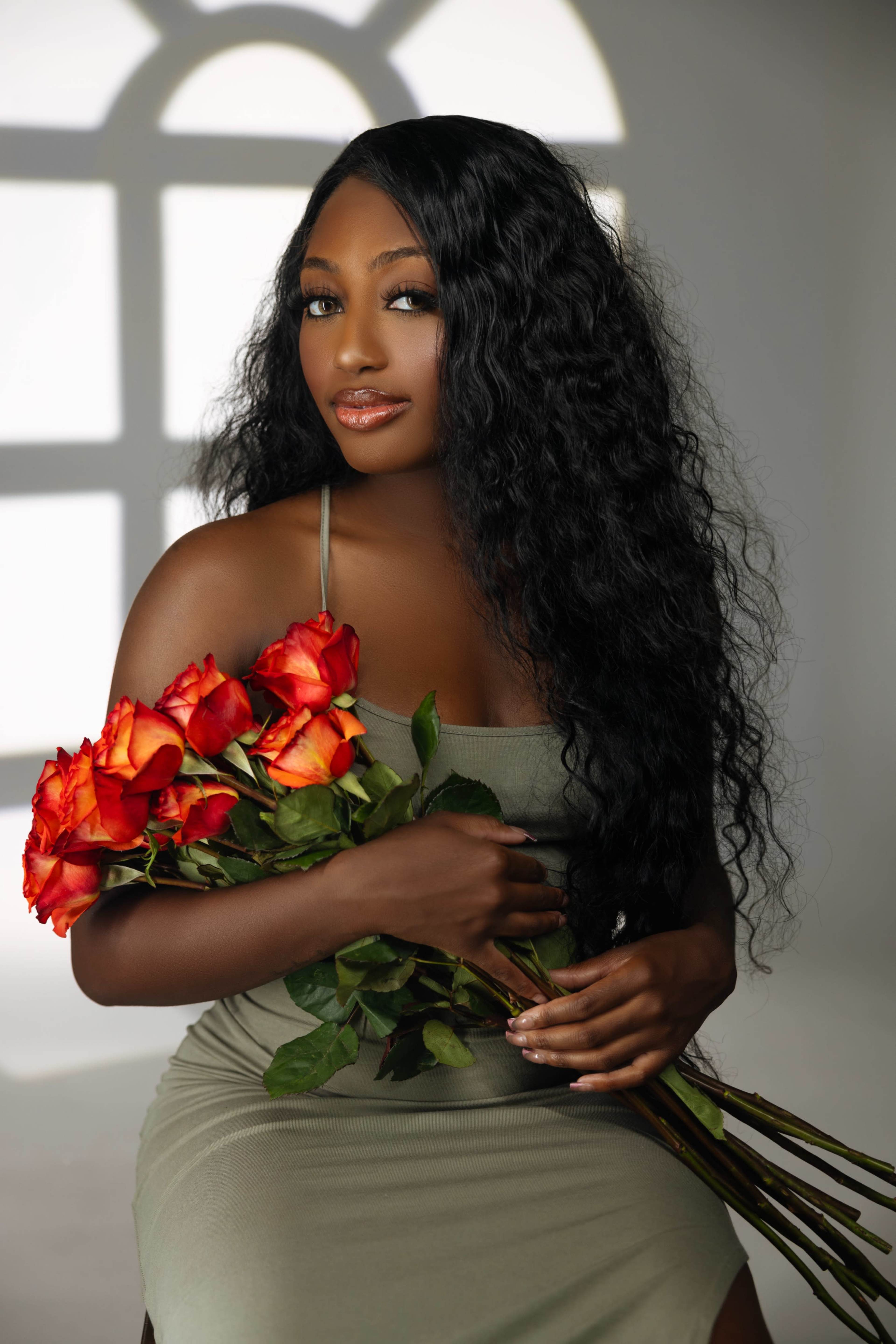 A woman with long, curly hair holds a bouquet of red and orange roses while seated in front of a large window.