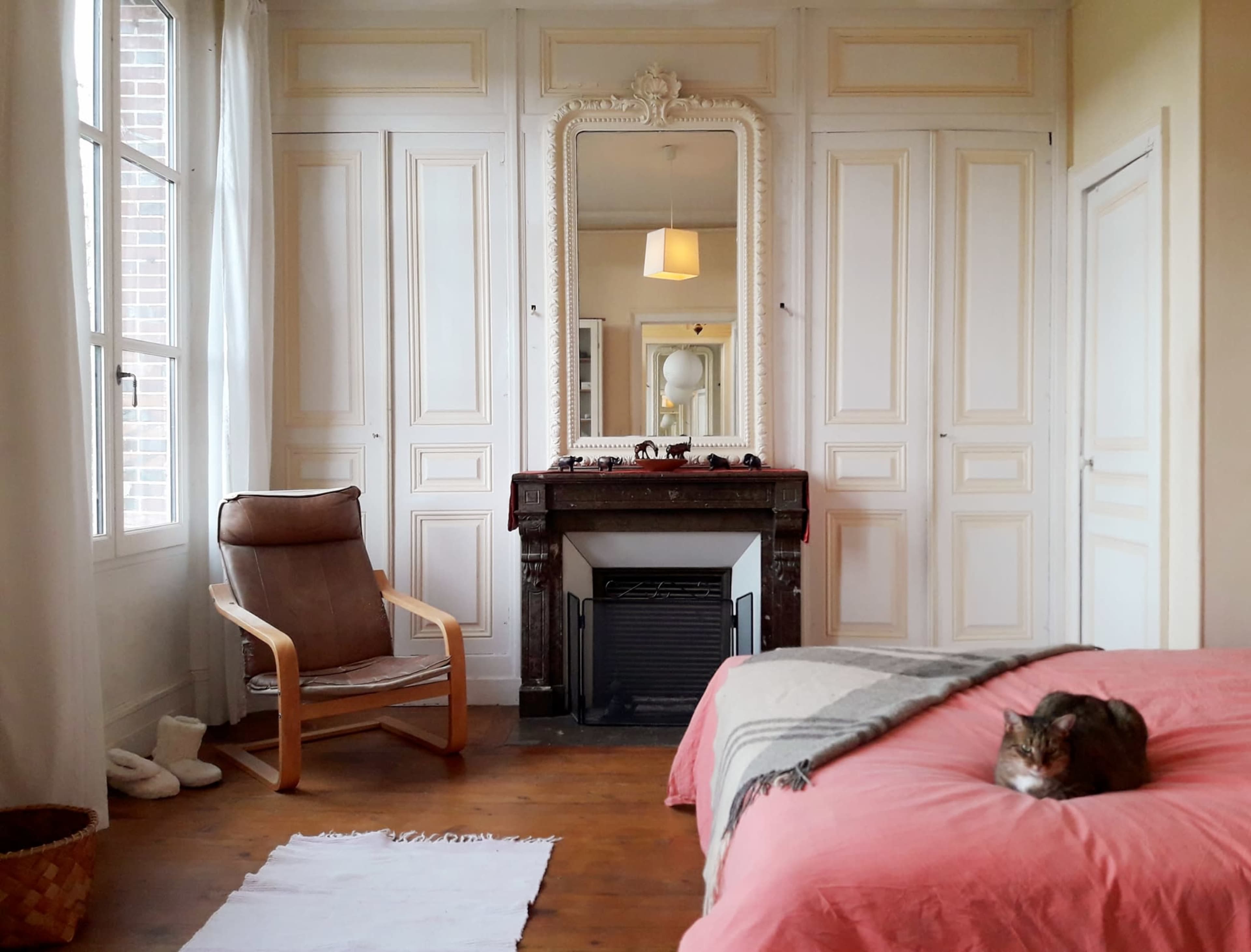 A cozy bedroom features a large mirror above a fireplace, a chair, and a cat resting on a pink bedspread.