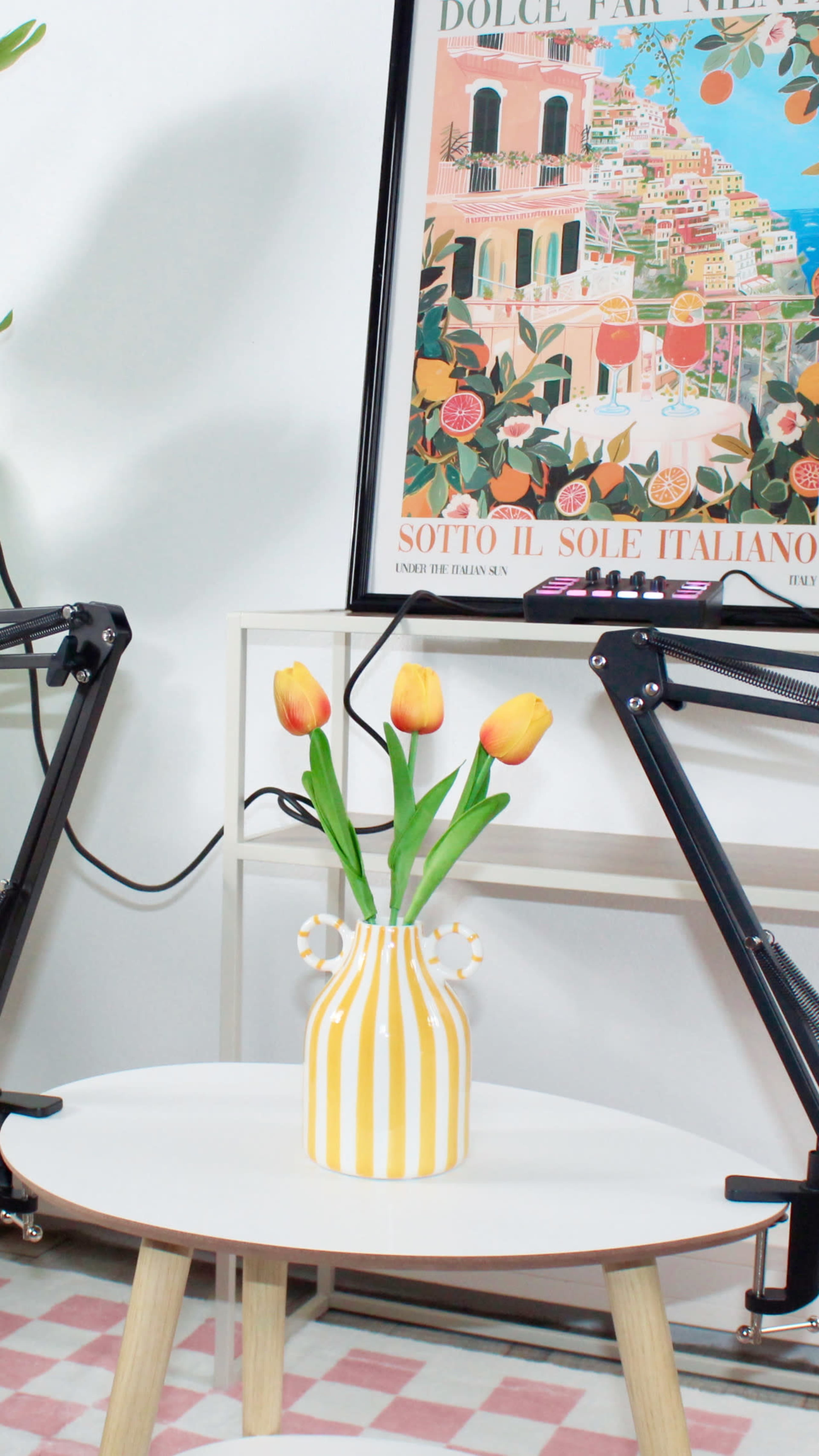 A yellow-striped vase with orange tulips sits on a round table near black adjustable arms and a colorful, framed poster on the wall.