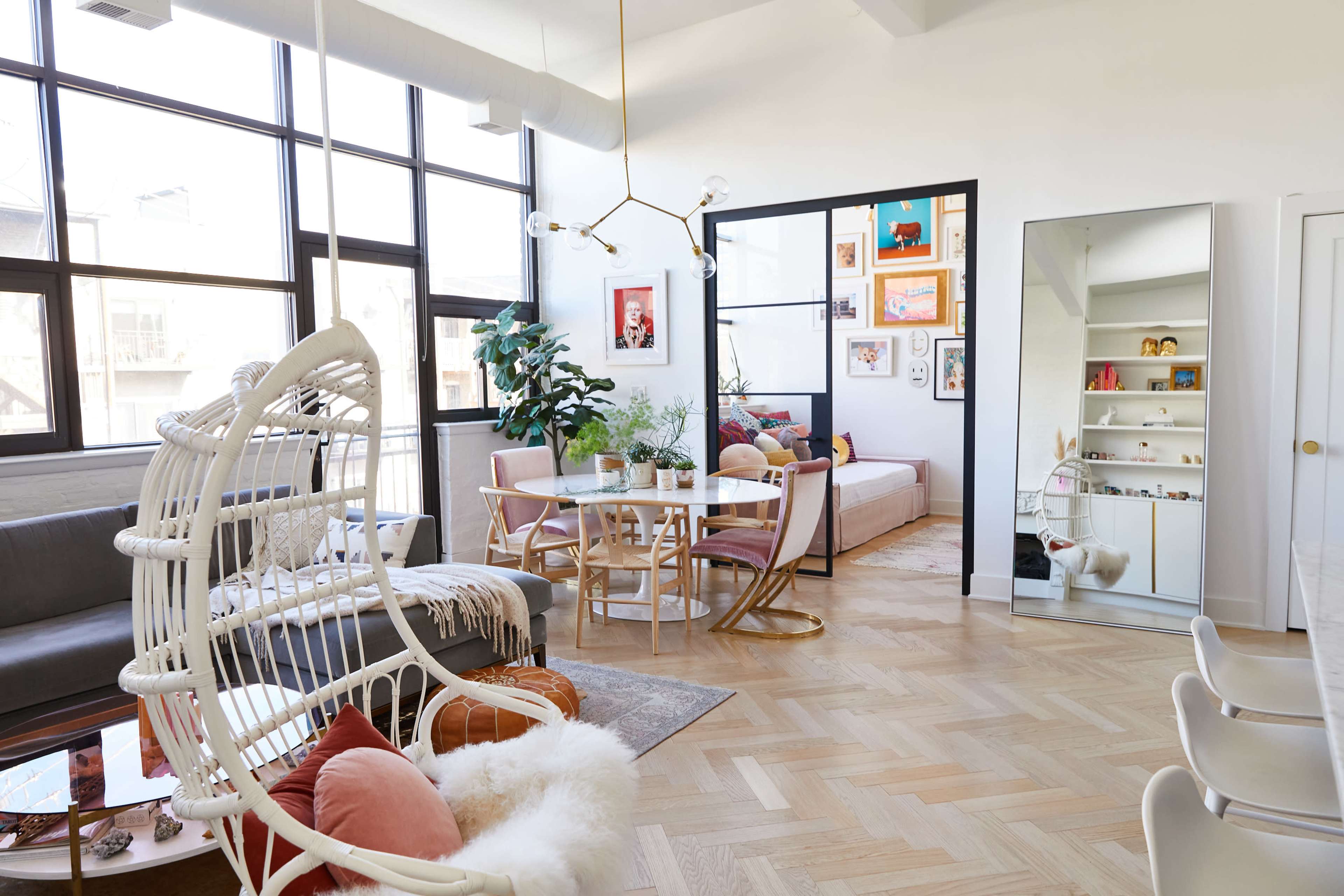 The image shows a stylish living area with a seating arrangement, a dining table, and large windows, featuring modern decor and plants.