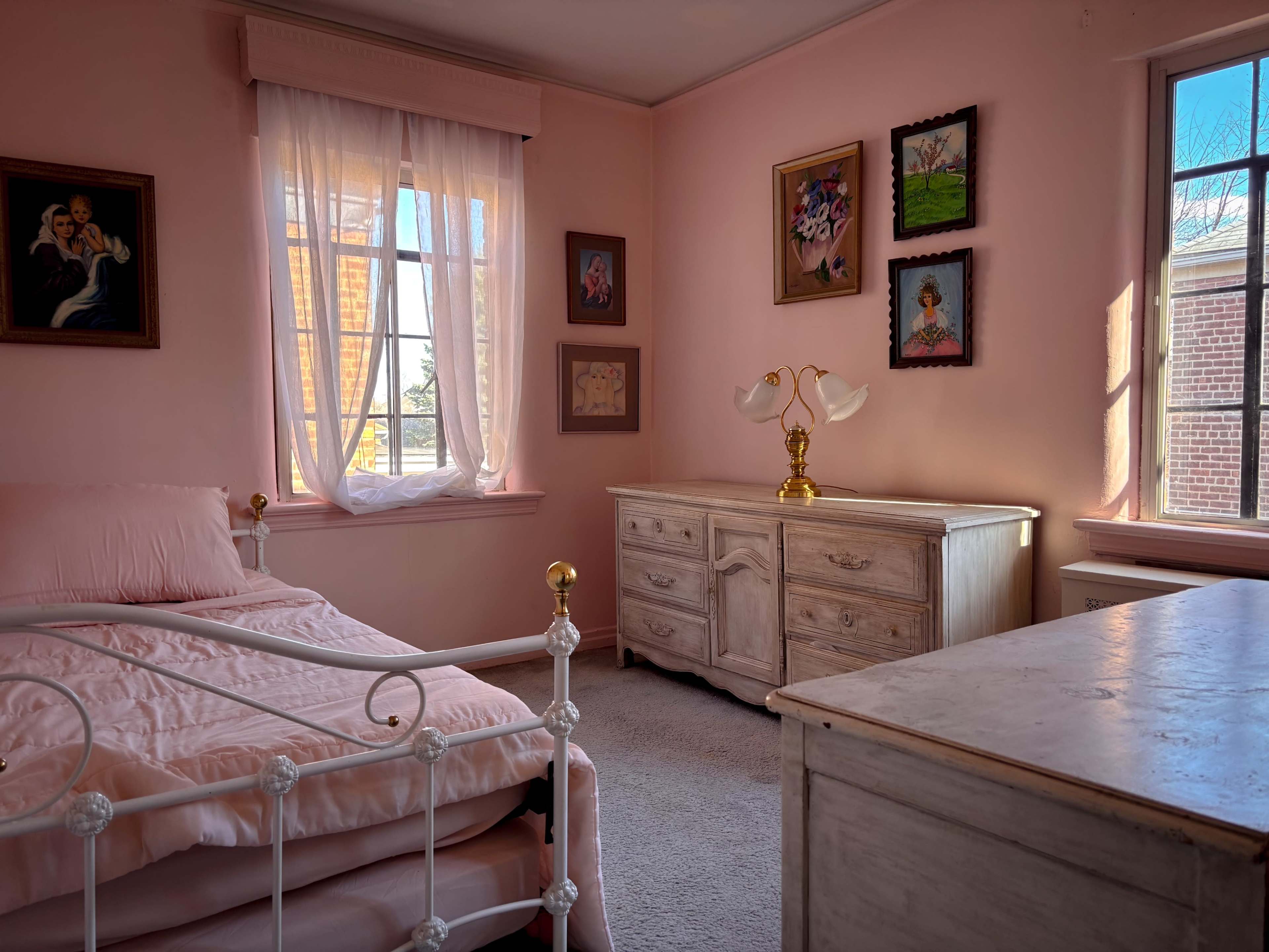 The image shows a softly-lit bedroom with a pink color scheme, featuring a bed, a dresser, and several framed artworks on the walls.