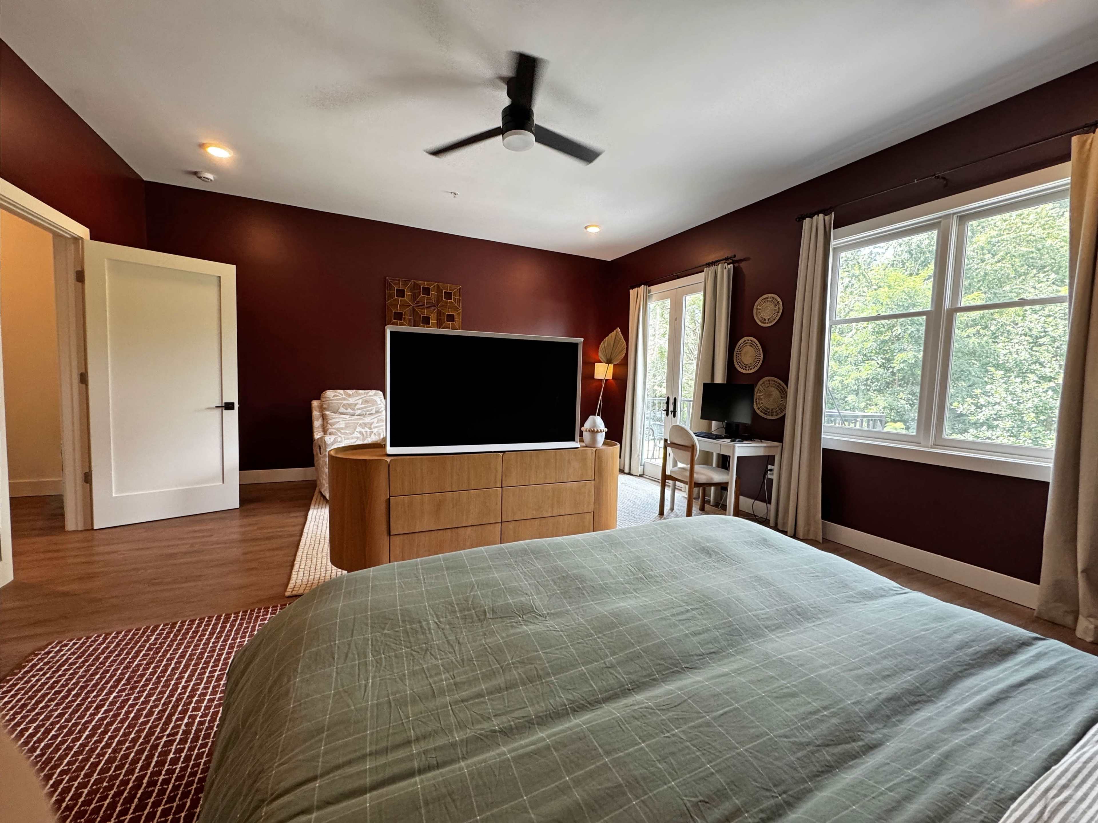 The image shows a bedroom with a dark red accent wall, a large bed, a television unit, and a small desk near a window.