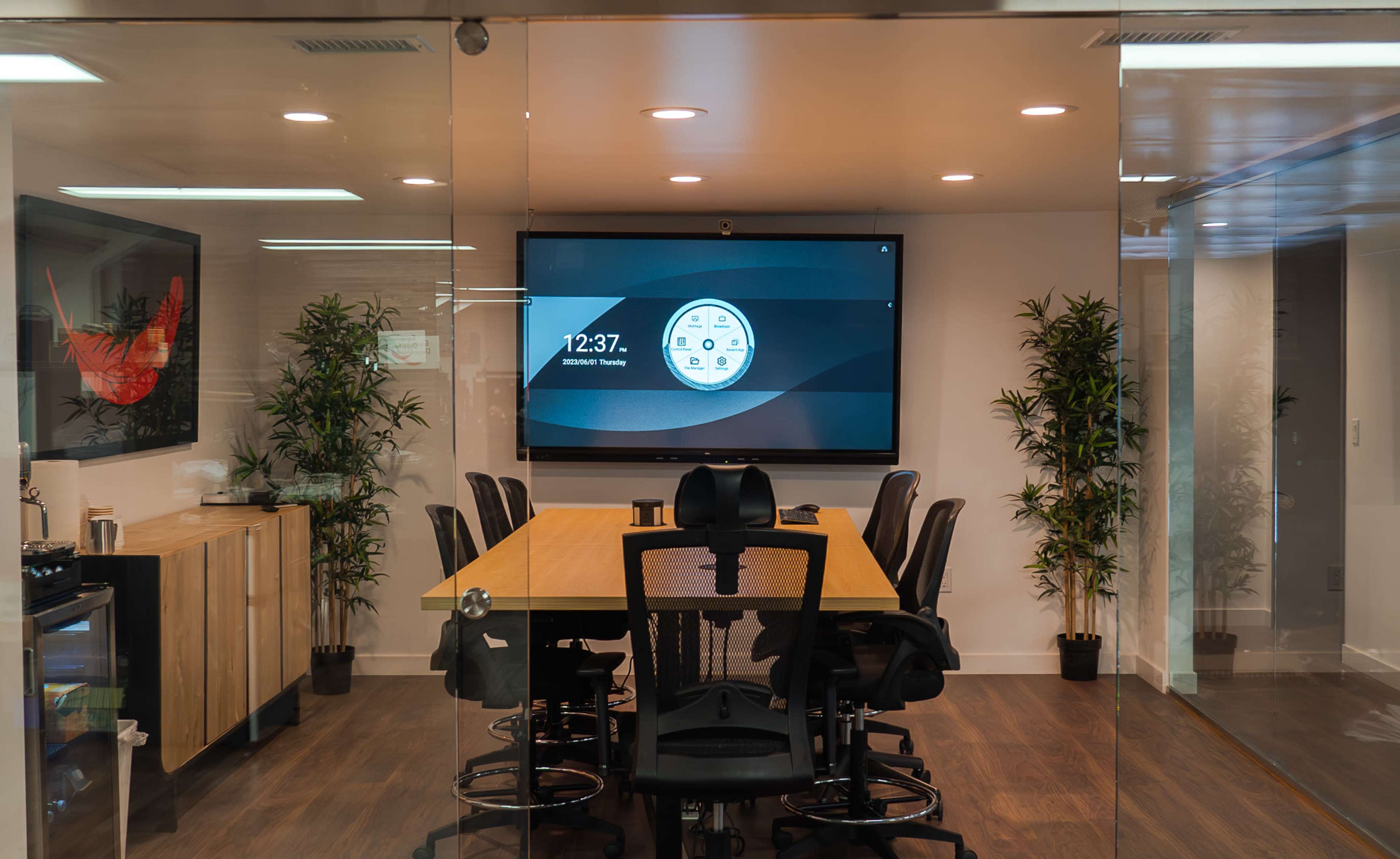 A modern conference room is visible through glass walls, featuring a long wooden table surrounded by black chairs and a large screen displaying the time.