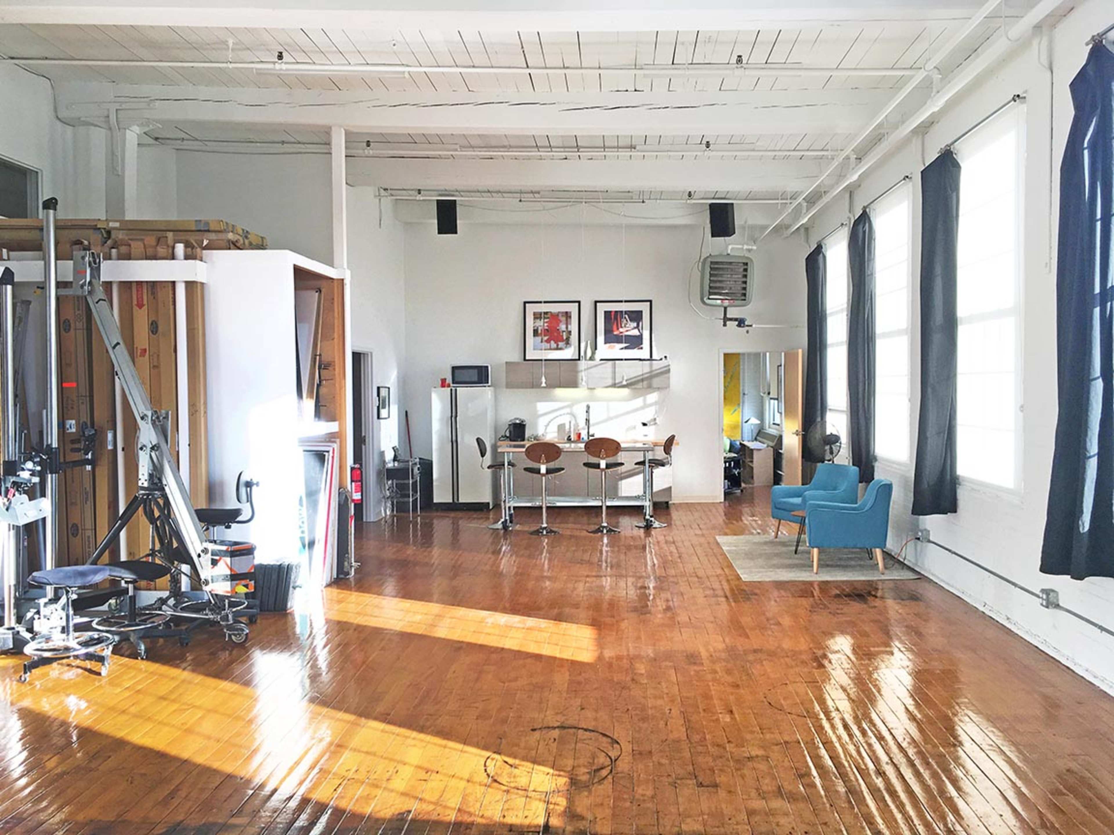 A spacious, well-lit room with wooden flooring, a fitness machine, a dining area with bar stools, and a cozy seating area featuring a blue chair.