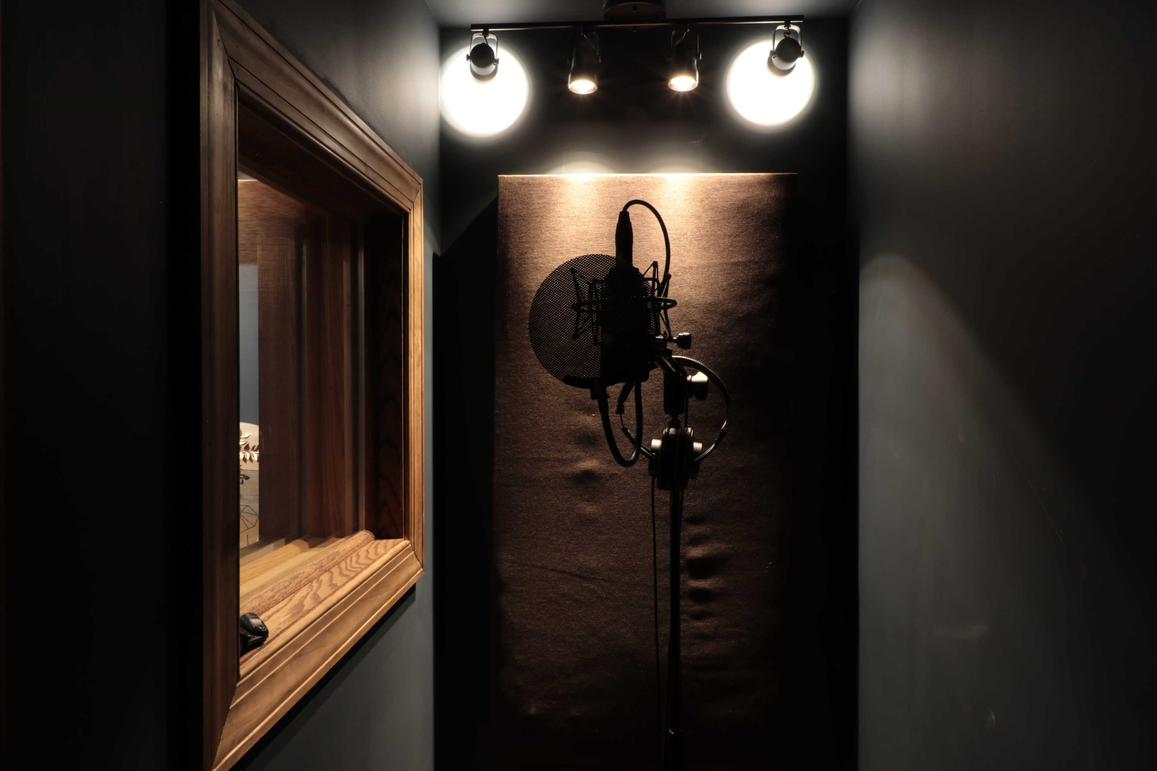 A dimly lit recording booth with a microphone on a stand in front of a sound-absorbing panel and a small window.