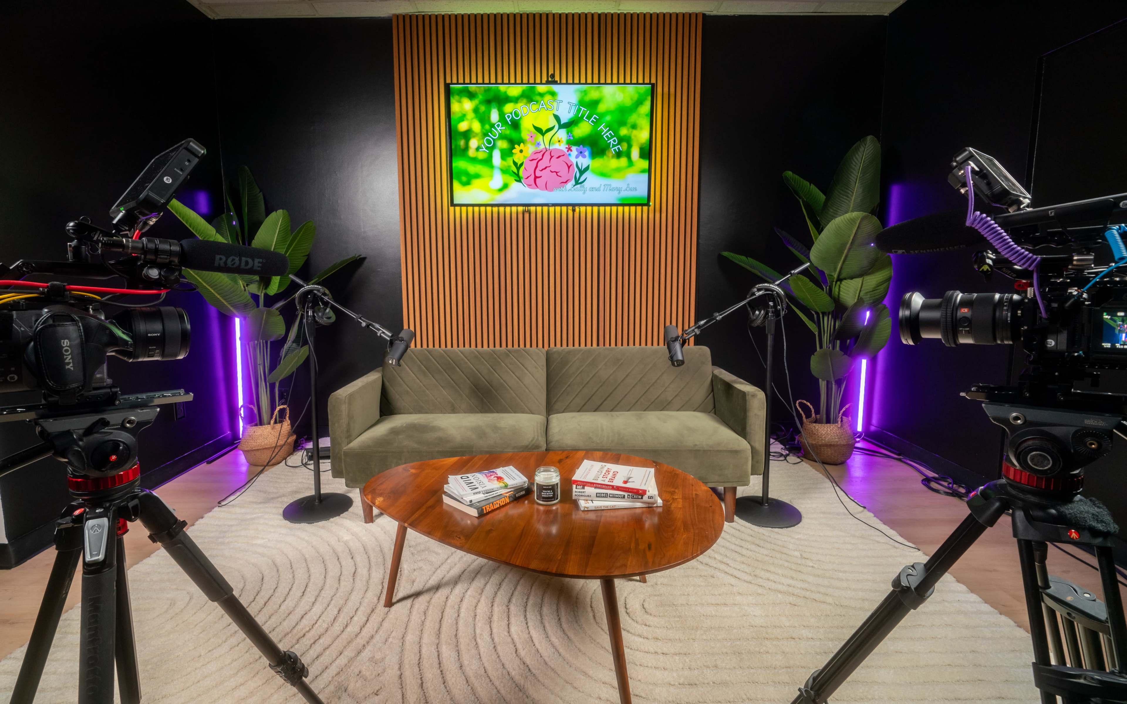 A video recording studio with a green sofa, a round wooden coffee table stacked with books, and two cameras on tripods facing a wall-mounted screen displaying a colorful logo.
