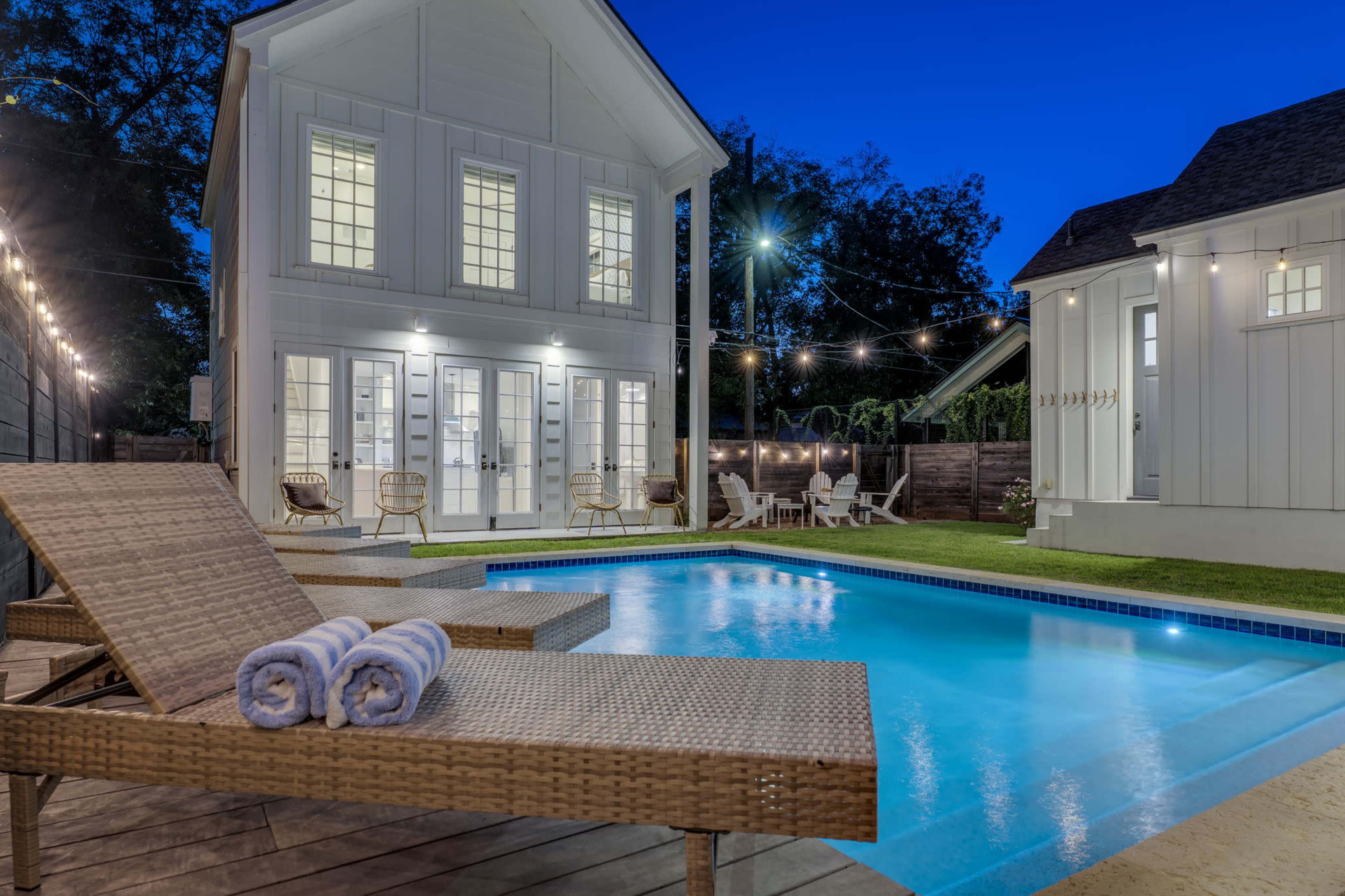 The image shows a modern house with a swimming pool and lounge chairs in a well-lit backyard during twilight.