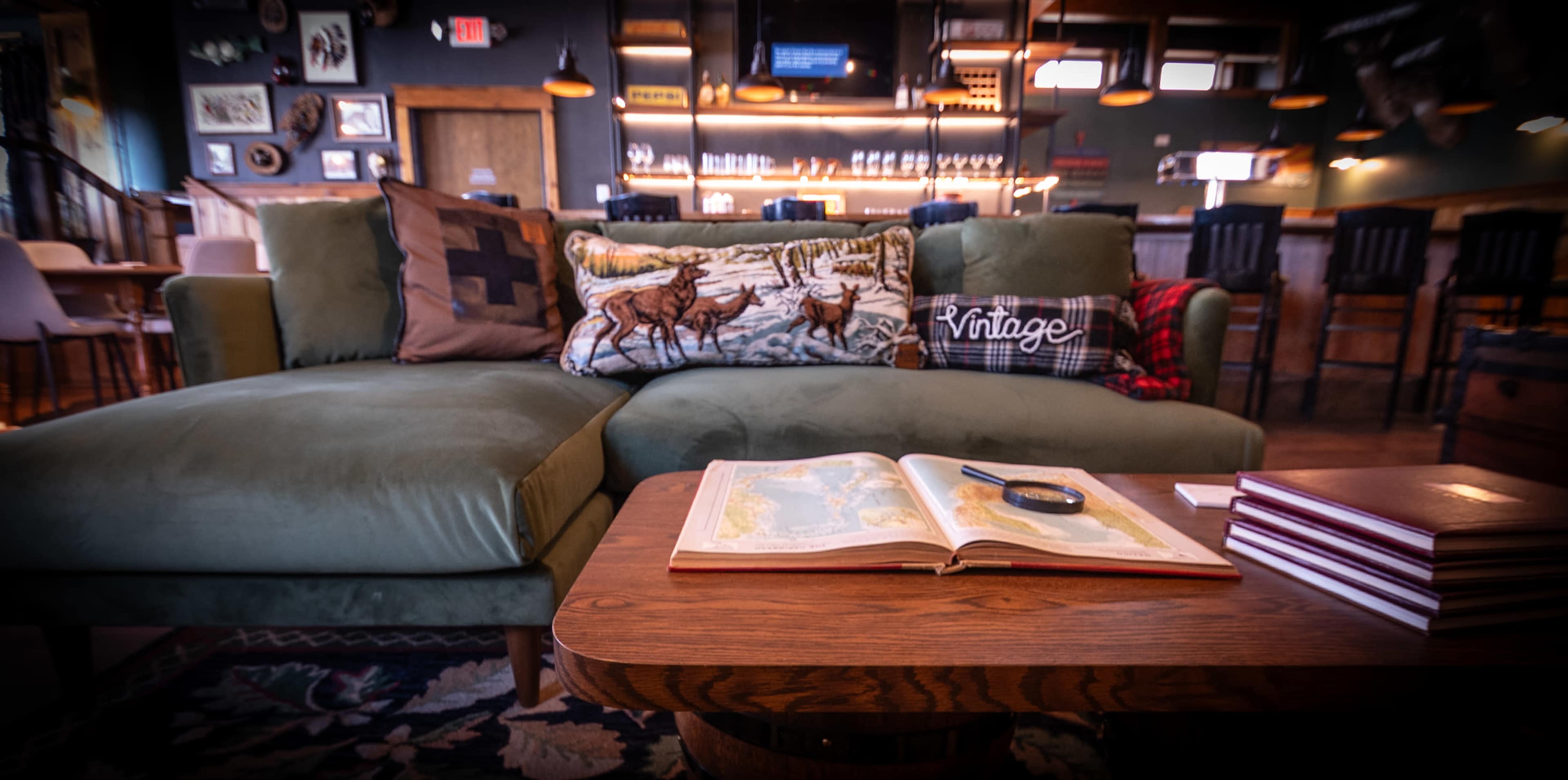The image features a cozy seating area with a green sectional couch adorned with decorative pillows, a wooden coffee table displaying an open book and a magnifying glass, set in a rustic bar environment.