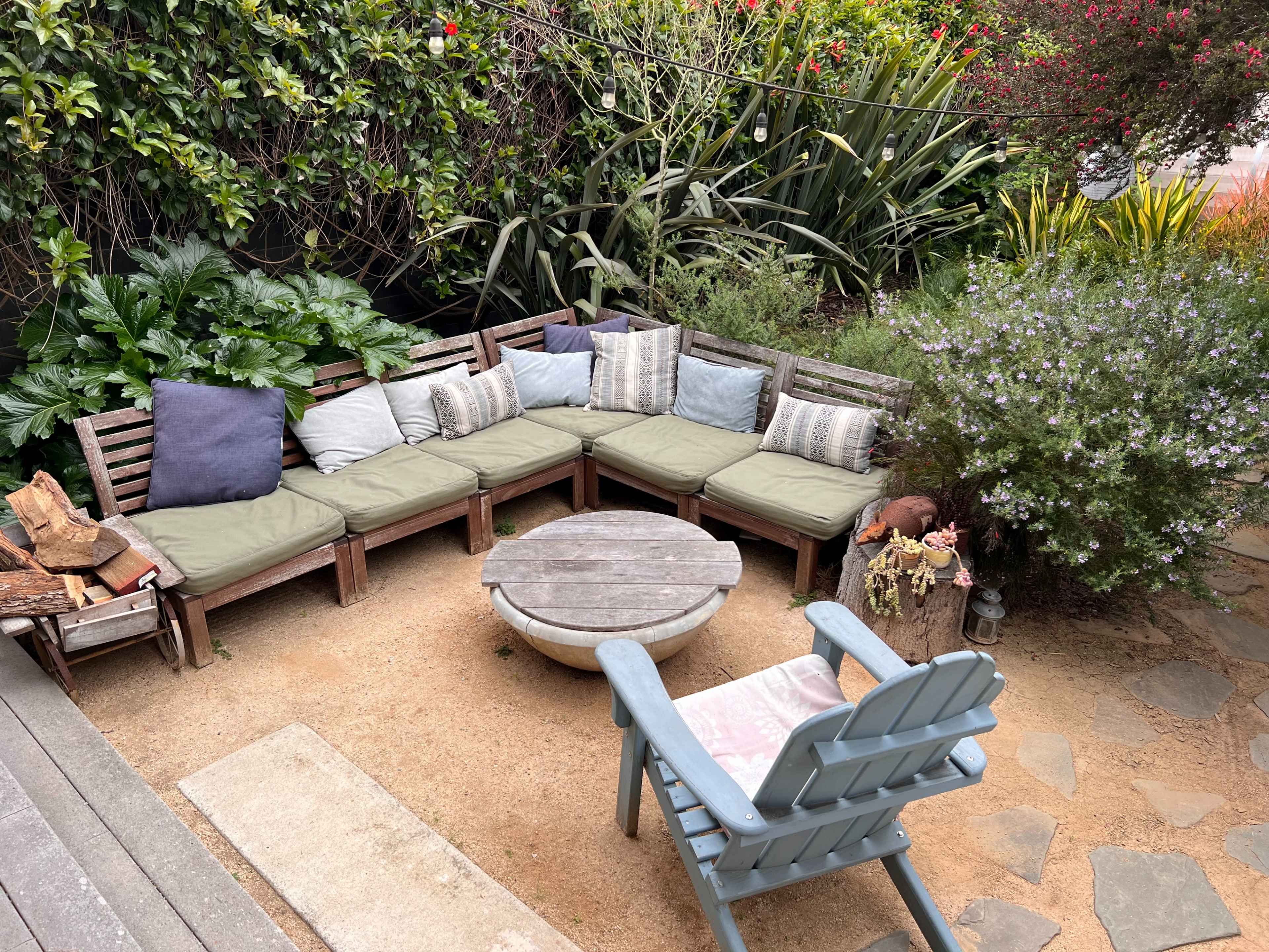 A cozy outdoor seating area features a wooden sectional sofa with cushions, surrounding a circular table and a fire pit, set amidst lush greenery and gravel.