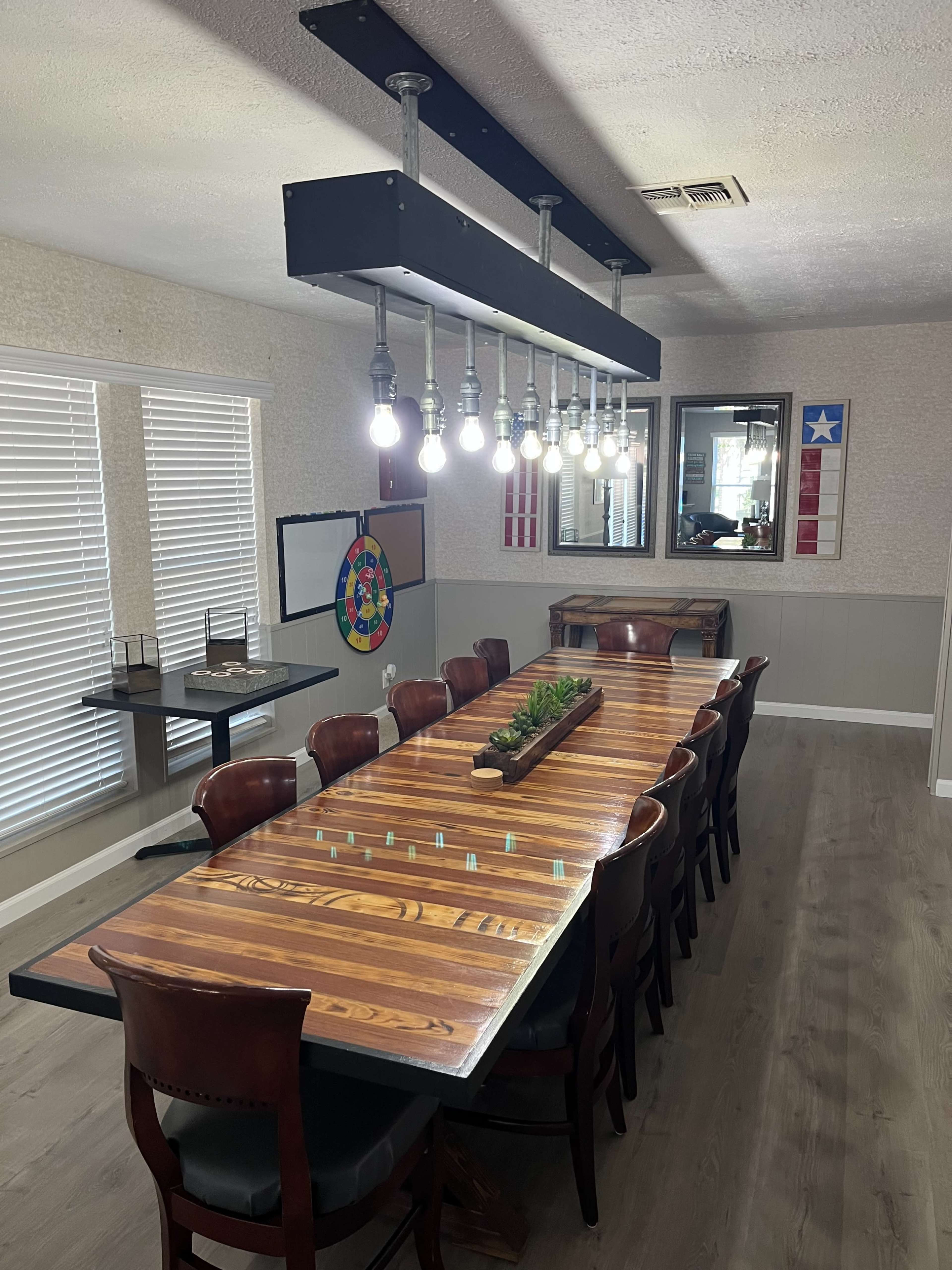 A long wooden dining table with eight chairs is illuminated by a row of hanging light bulbs, surrounded by walls adorned with a mirror and Texas-themed decor.