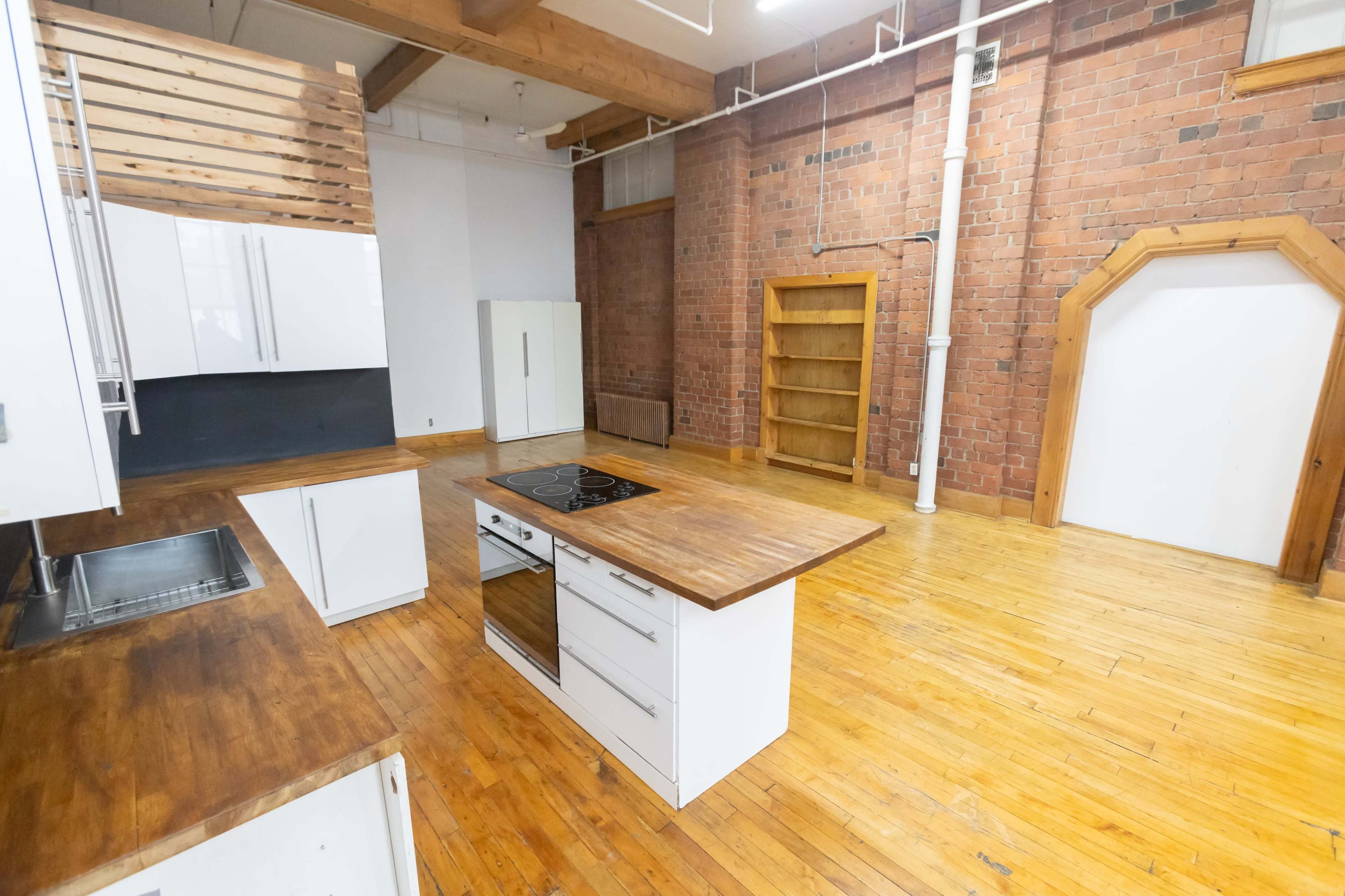 The image shows a modern kitchen with wooden countertops, white cabinets, and exposed brick walls, featuring a central cooking island and a bookshelf against the back wall.