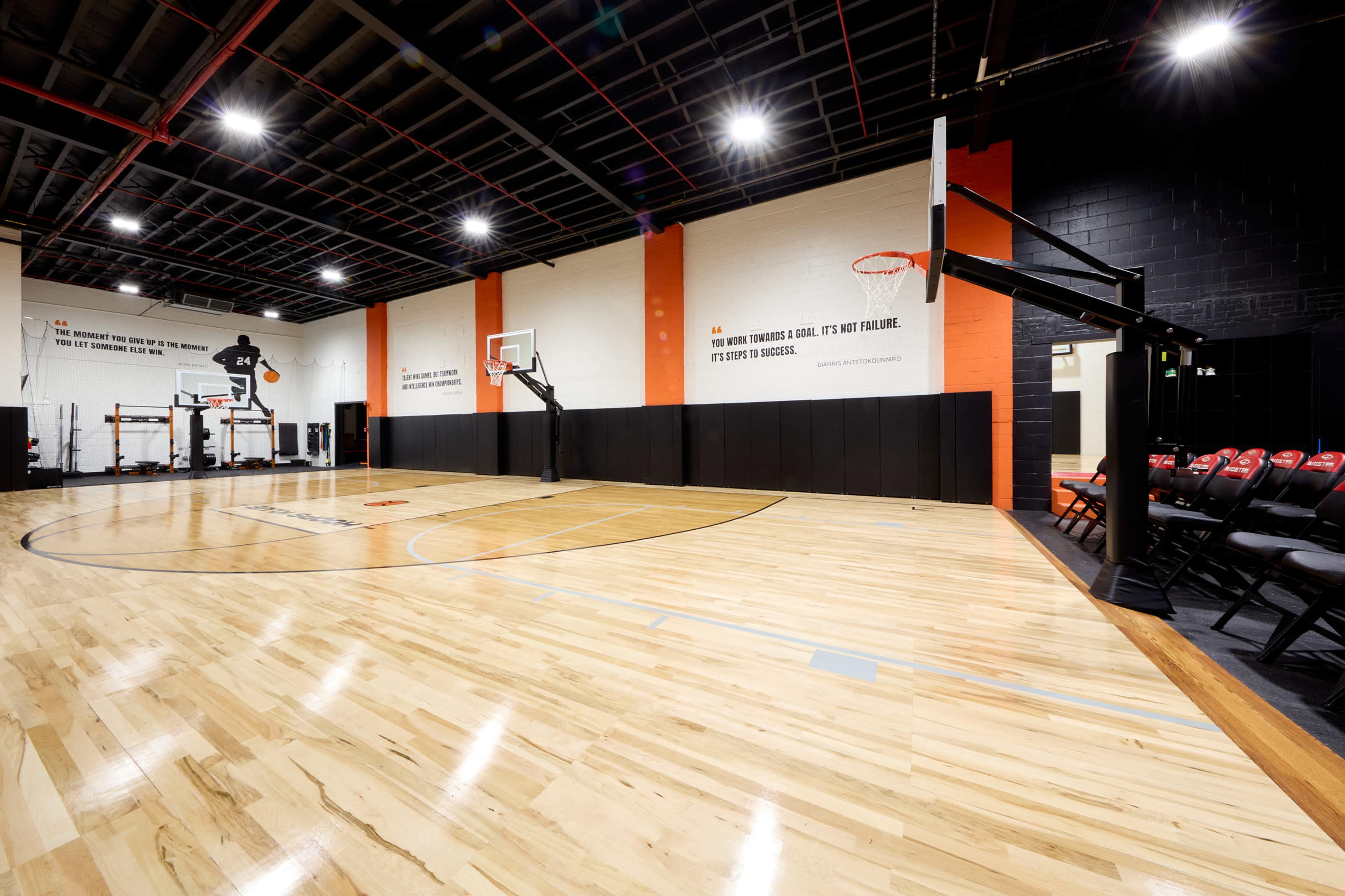 The image shows a modern indoor basketball court with wooden flooring, two basketball hoops, and padded wall protection, accompanied by motivational wall art.