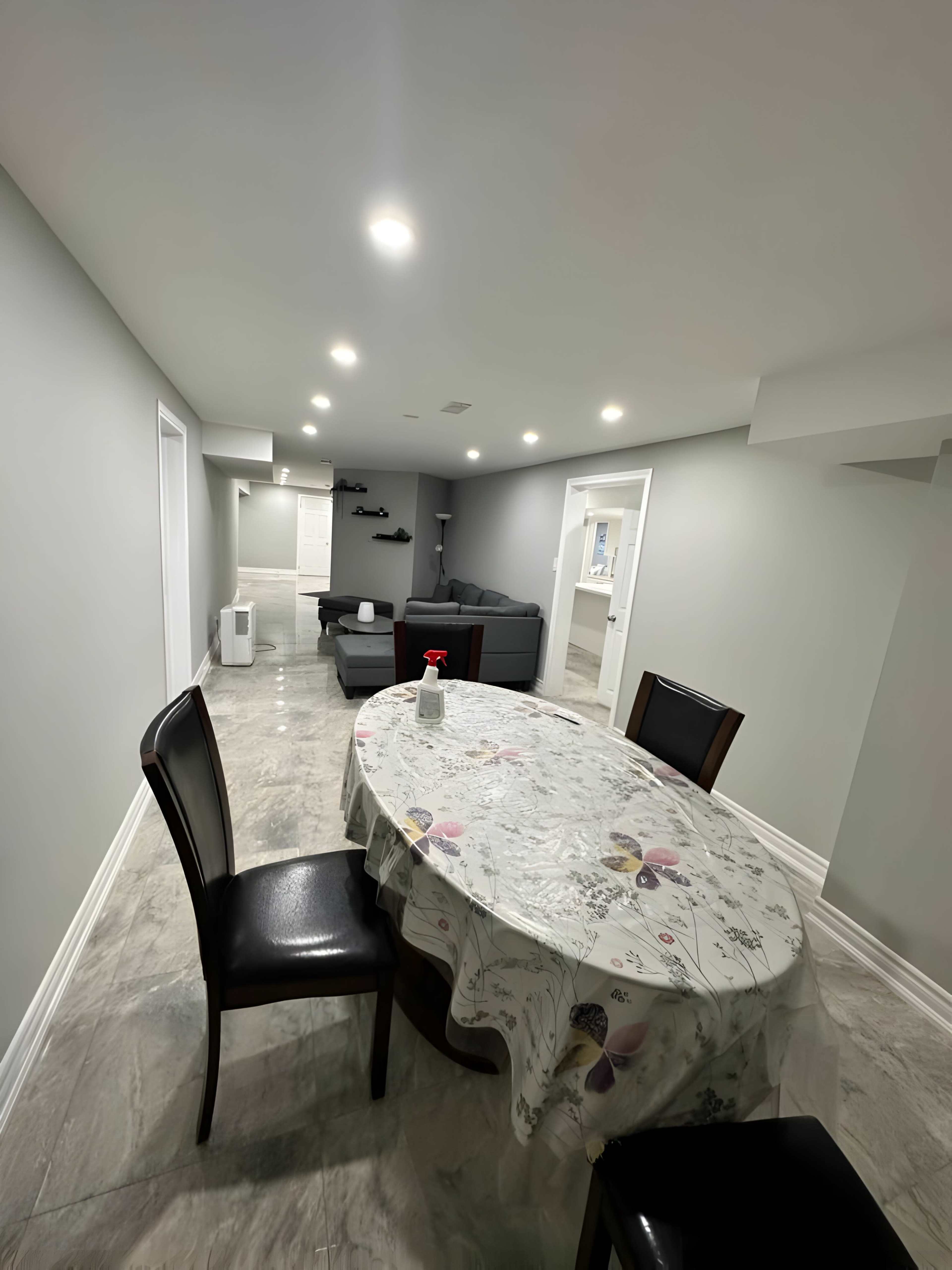 A modern basement area features a dining table with a floral tablecloth, four chairs, and a seating area with a couch in the background.
