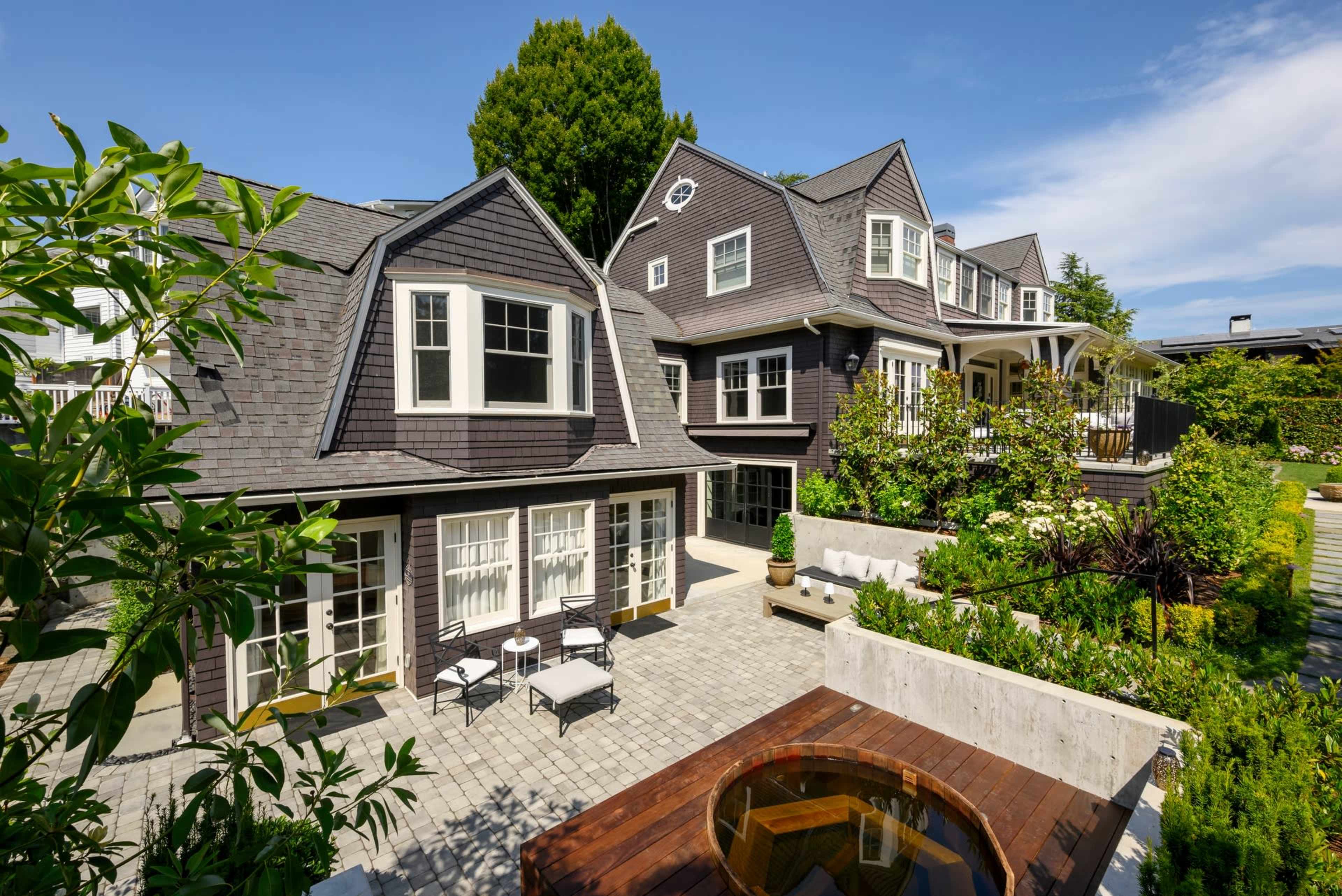 A two-story house with a gray shingle roof, surrounded by a landscaped yard featuring a patio, seating area, and a hot tub.