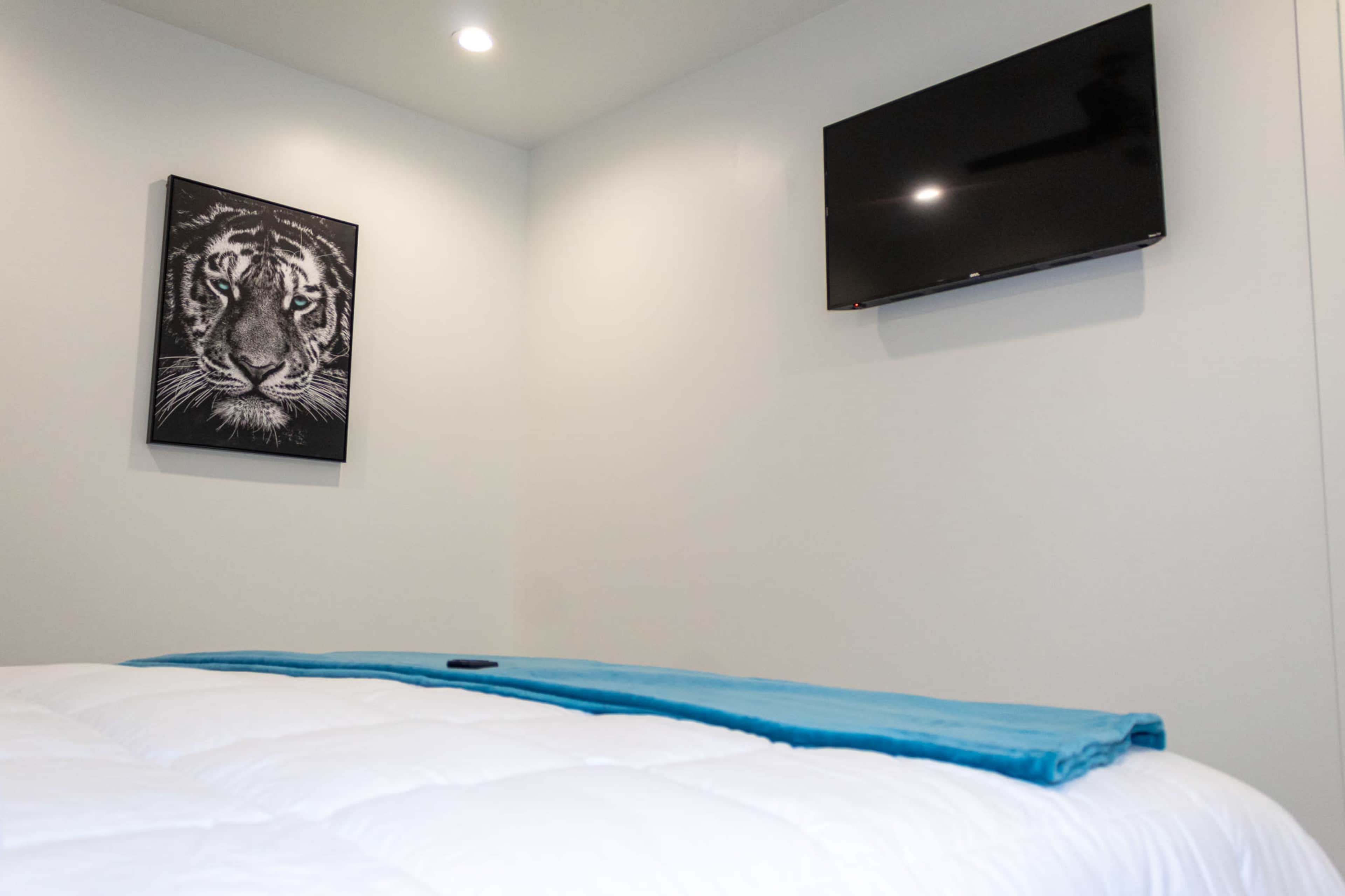 The image shows a bedroom with a white bedspread, a blue blanket draped across the bed, a wall-mounted television, and a framed tiger portrait on the wall.