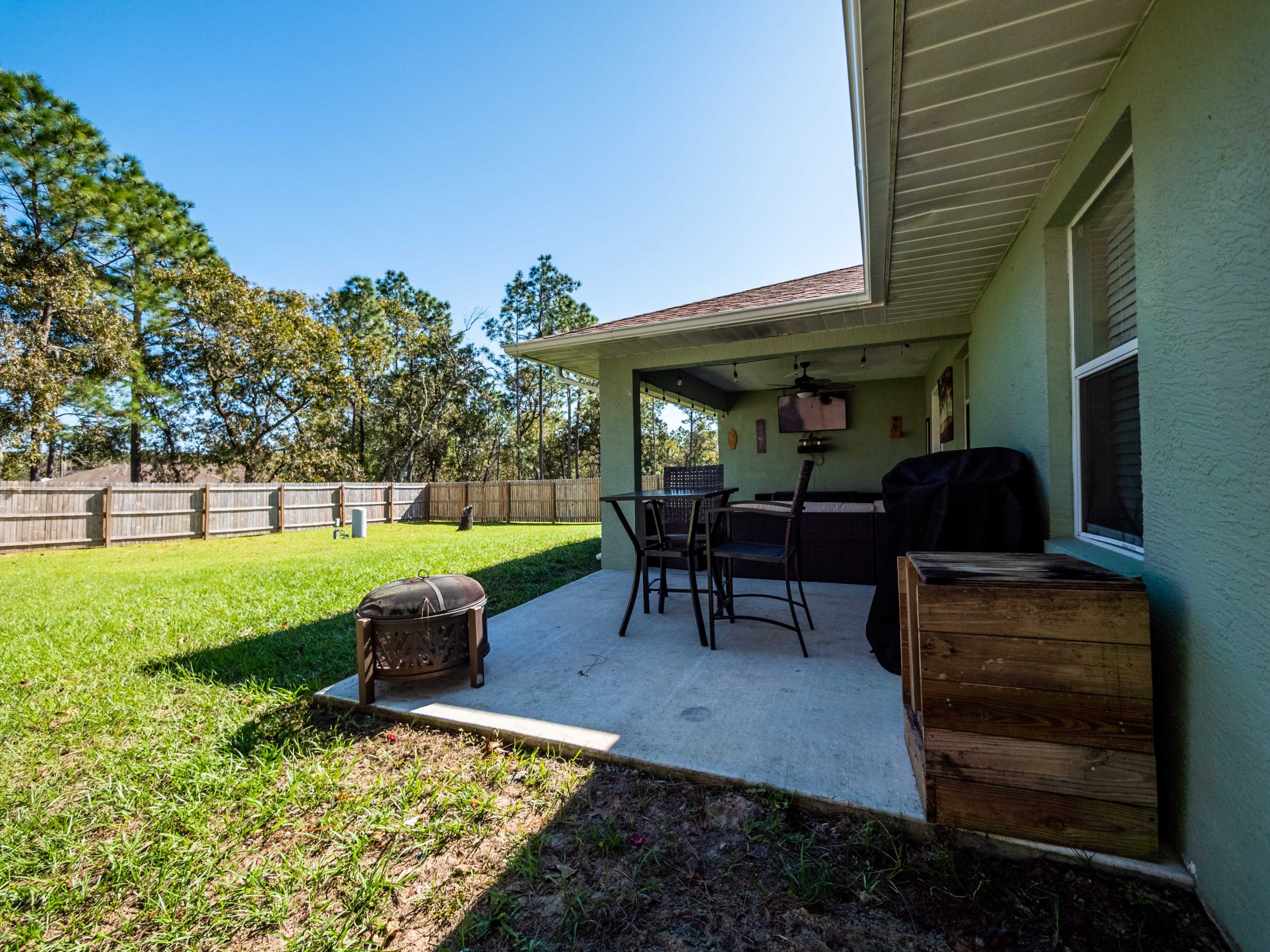 A patio area with outdoor seating, a charcoal grill, and a view of a grassy backyard enclosed by a wooden fence.