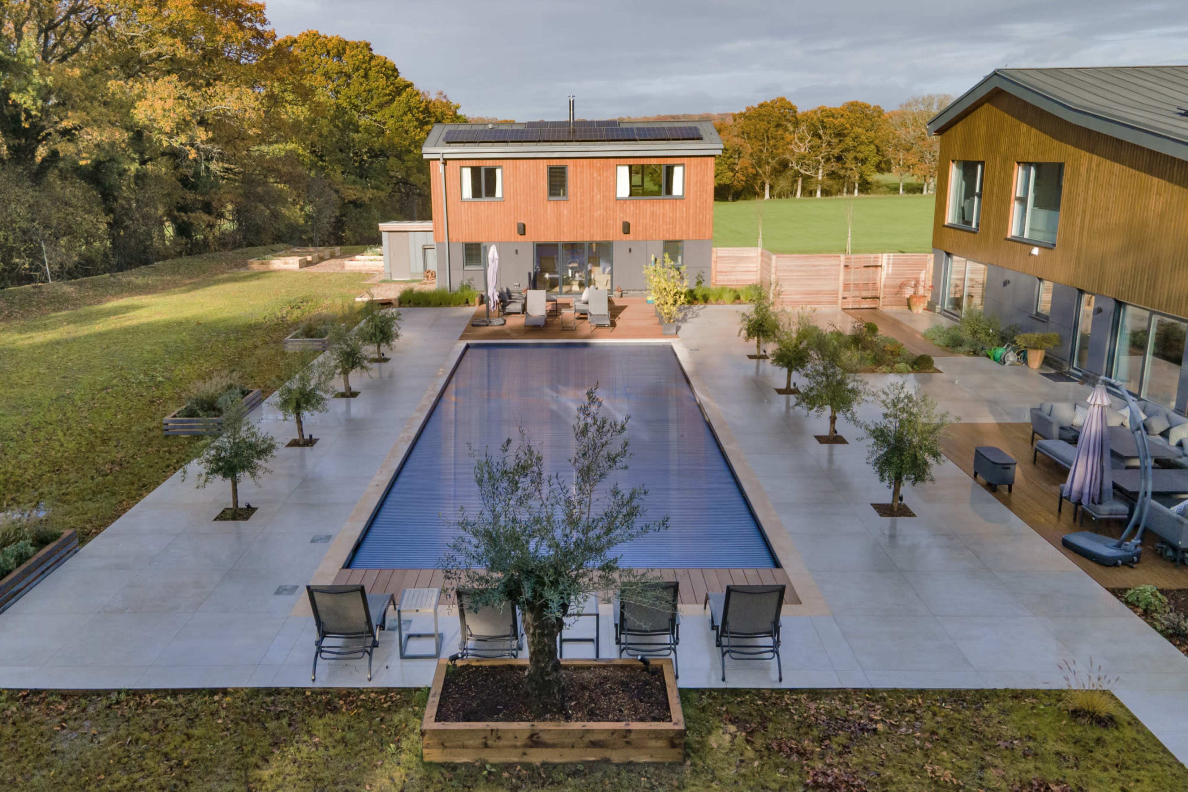 The image shows a modern outdoor area featuring a large swimming pool surrounded by landscaped trees, lounge chairs, and two buildings with wooden facades.