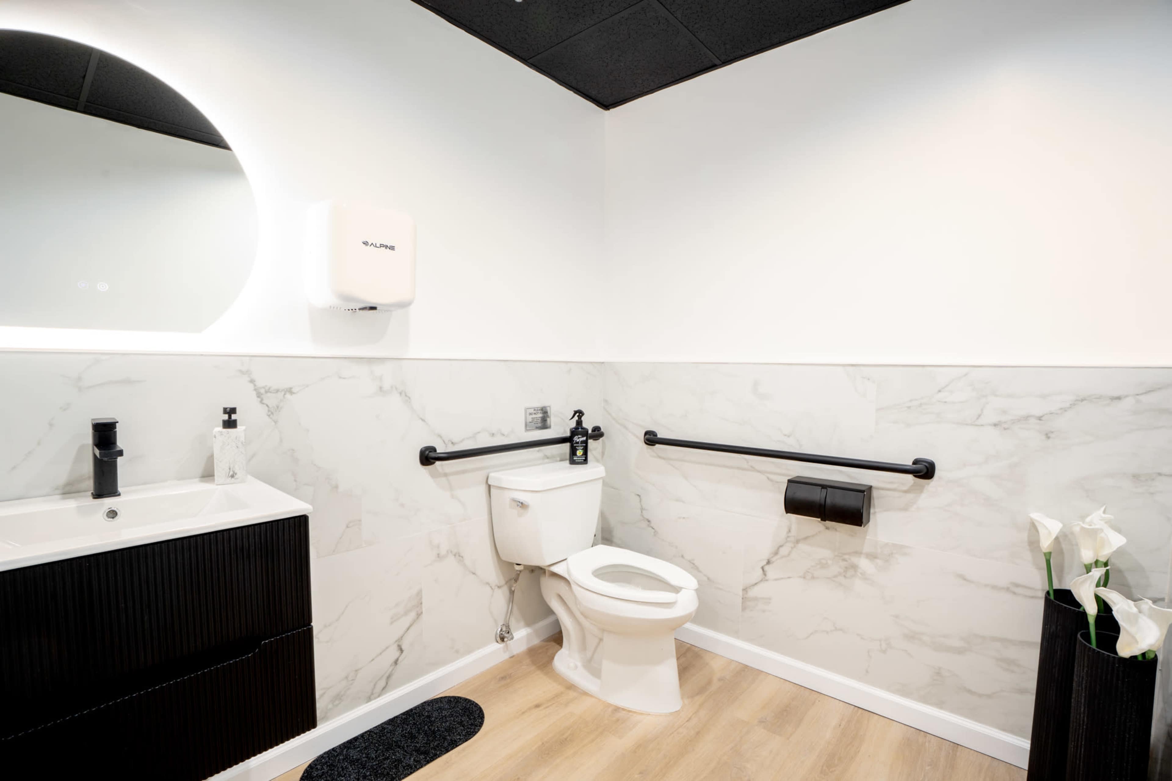 The image shows a modern restroom featuring a white toilet, a sleek sink with a black cabinet, and marble-style wall panels, along with grab bars and a black toilet paper holder.