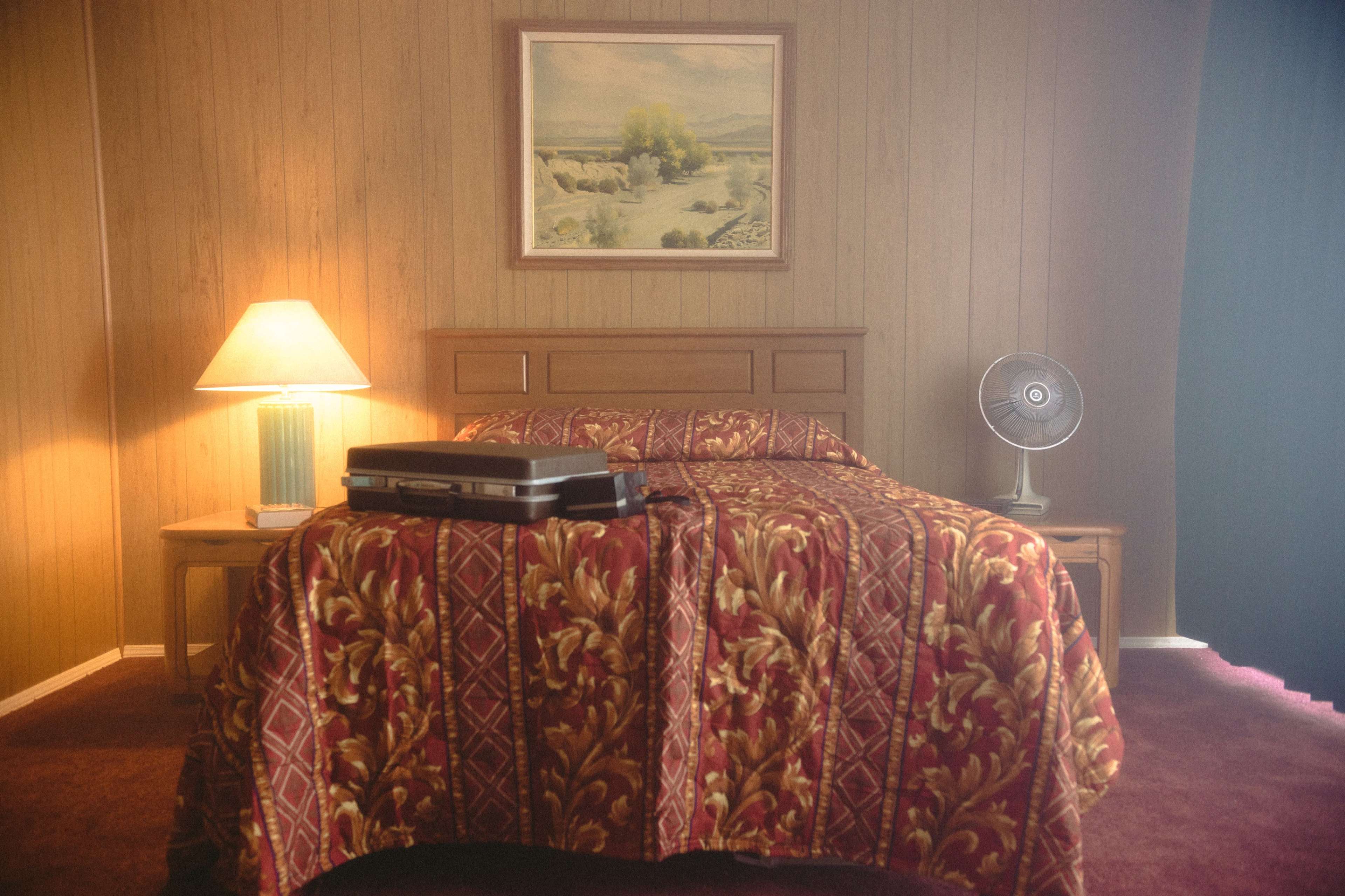 A bedroom features a bed with a patterned red bedspread, a wooden nightstand, a lamp, a fan, and a painting of a landscape on the wall.