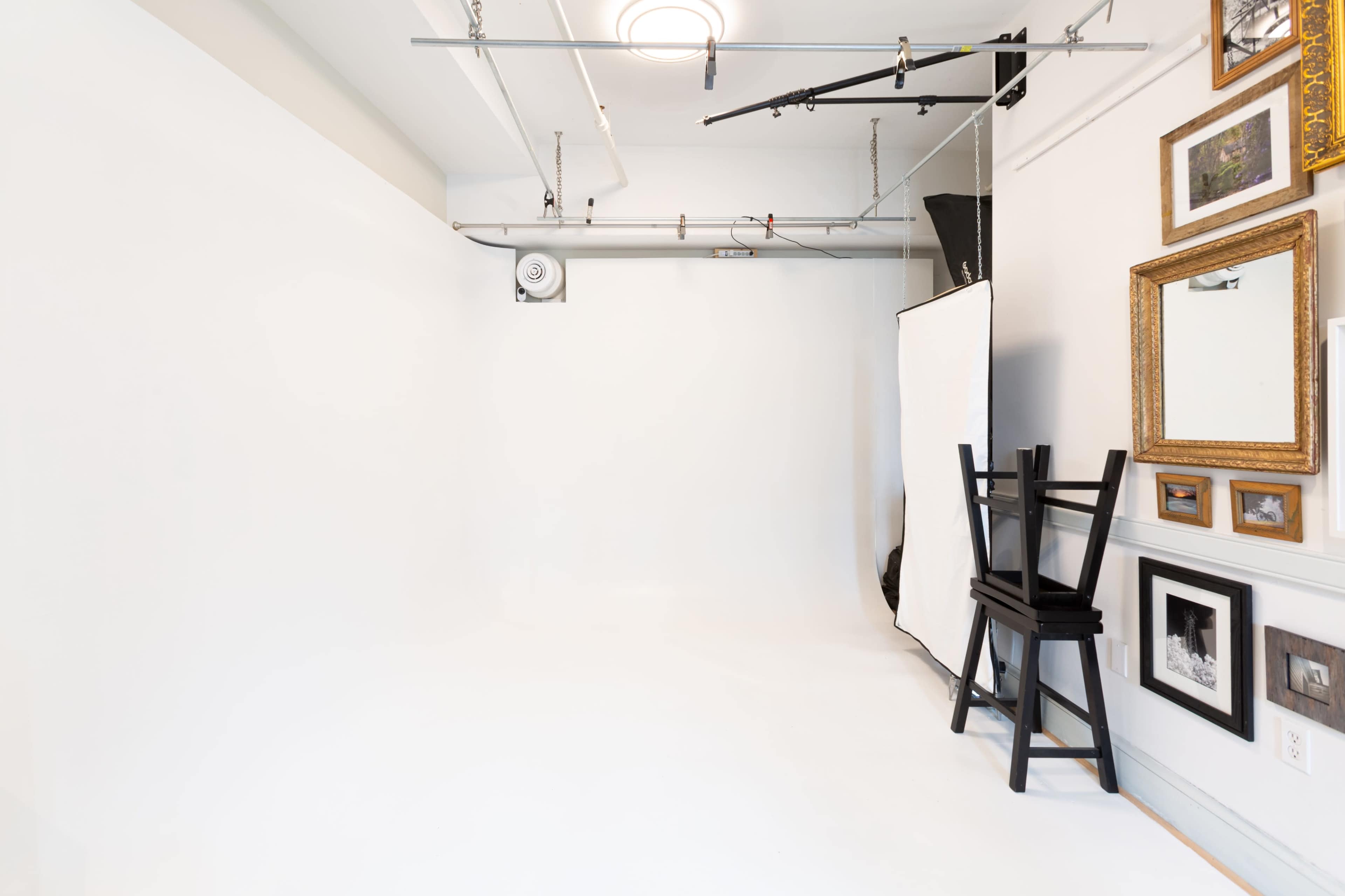 A photography studio features a blank white backdrop, a black chair, and various framed pictures hung on the wall.
