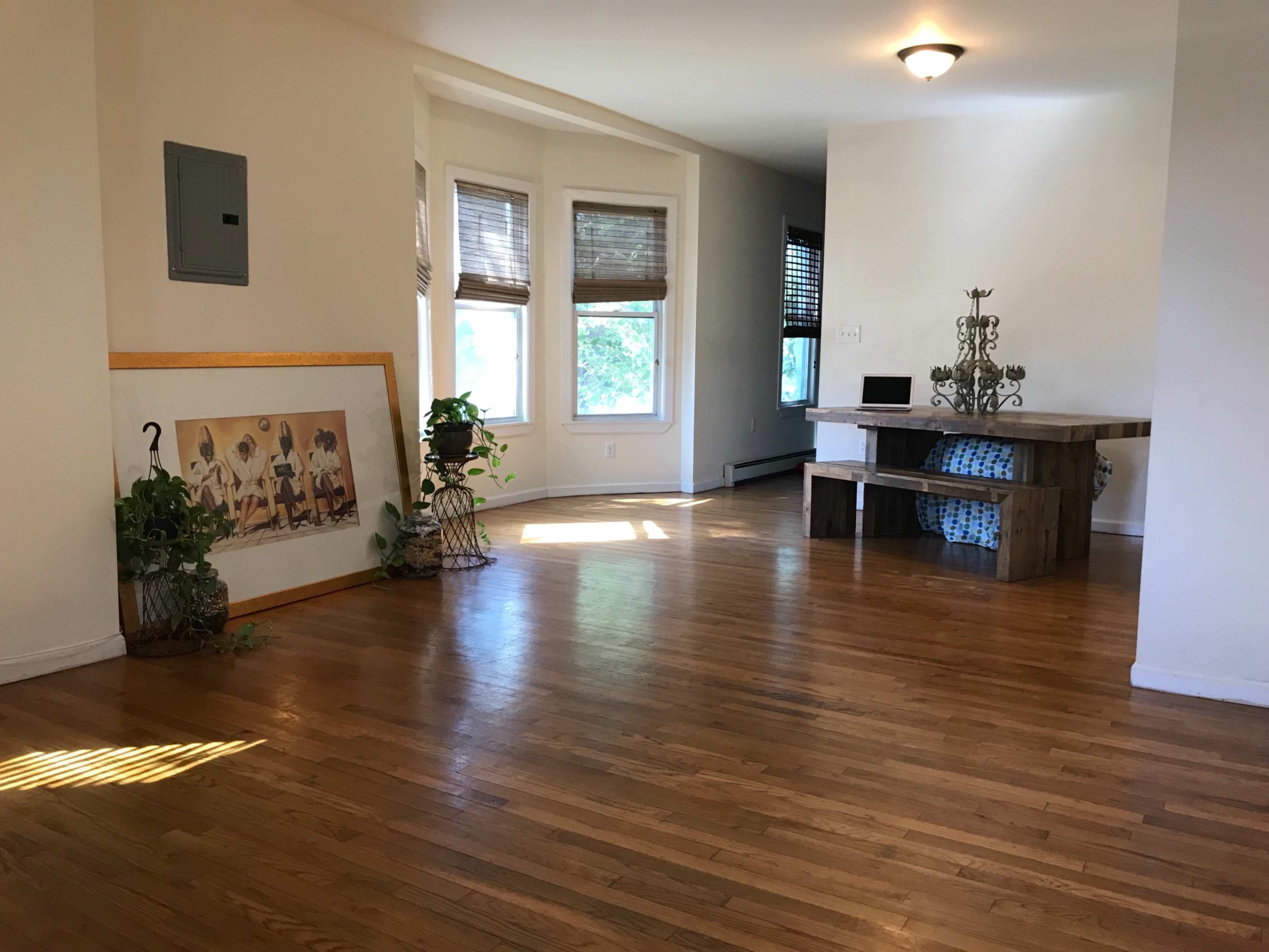 The image shows an interior space with hardwood floors, a large framed artwork leaning against the wall, and a wooden table in a well-lit area near windows.