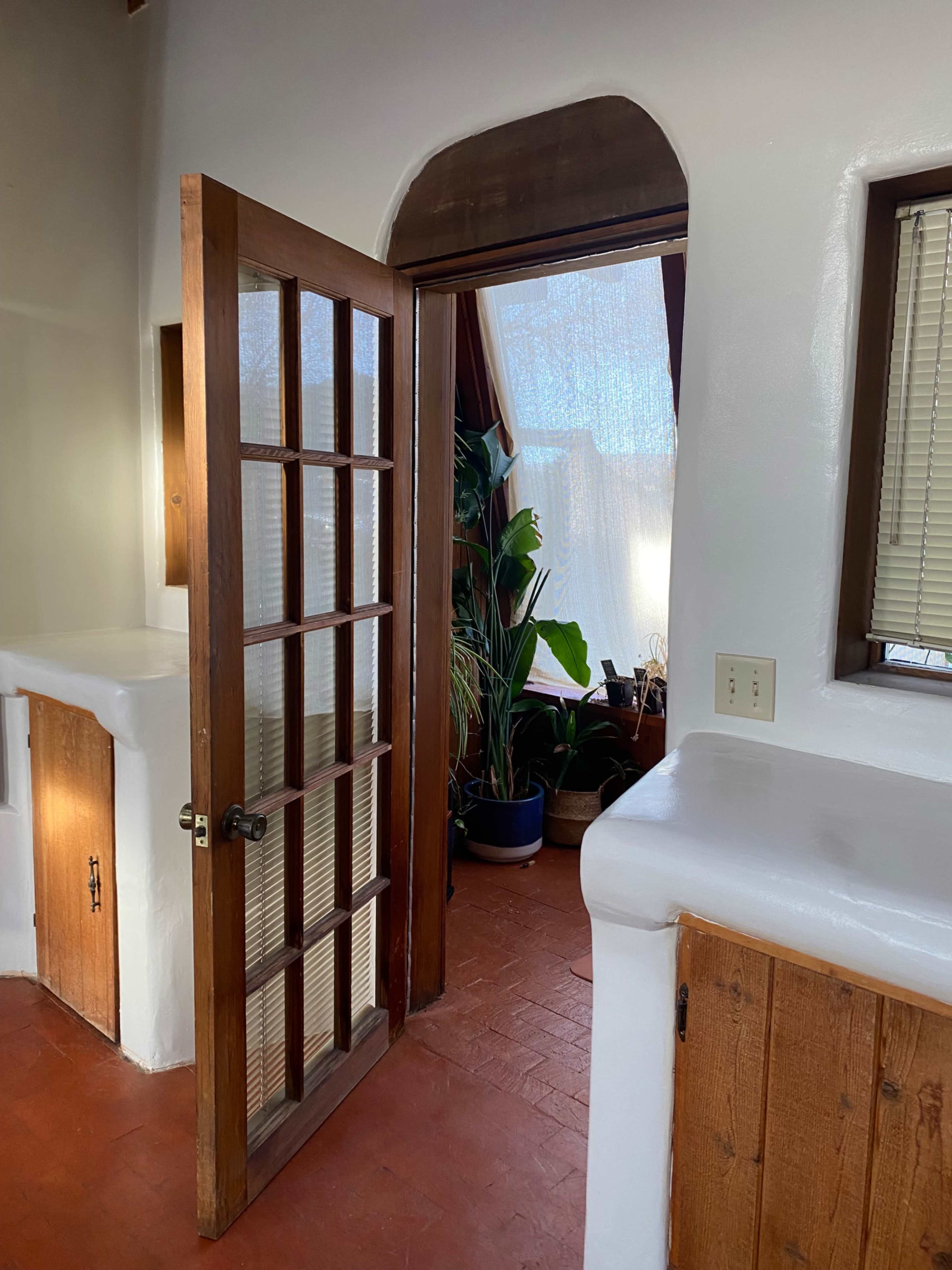 A wooden door with glass panels opens to a sunlit room featuring plants and beige walls.