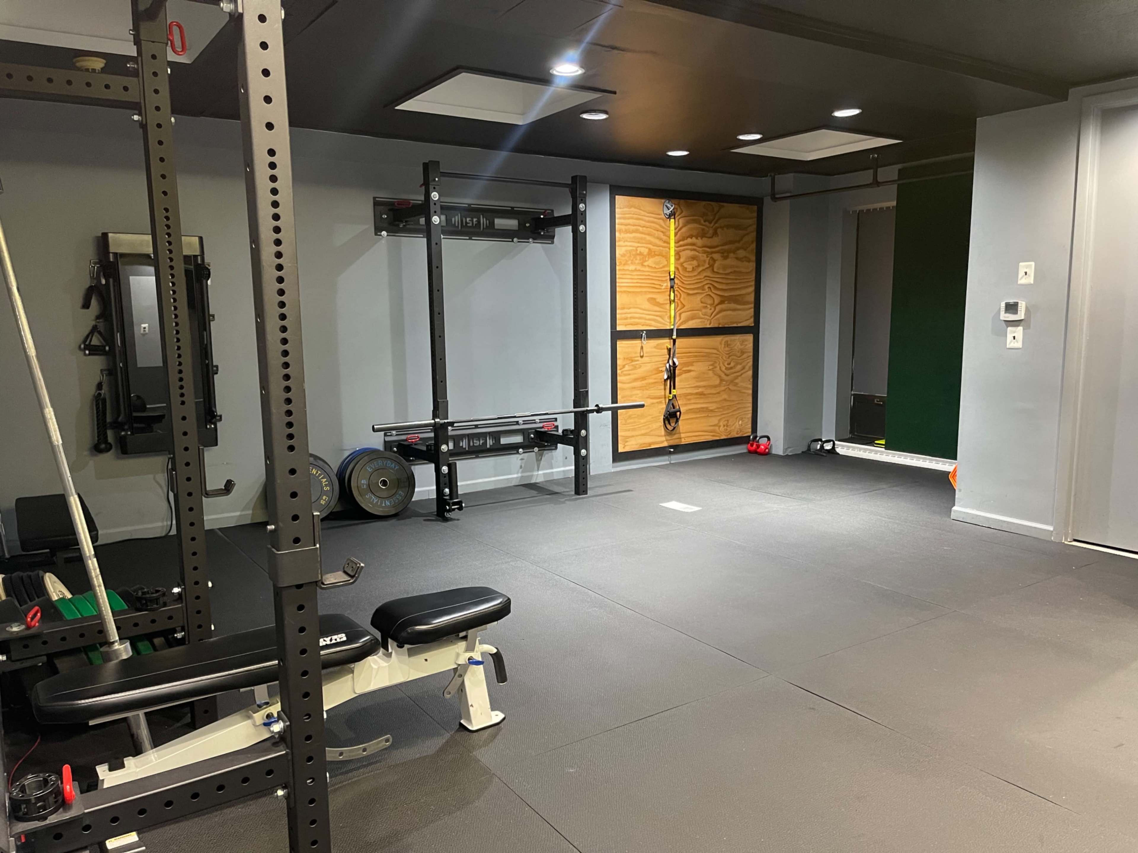A gym interior featuring a weight bench, squat rack, and wall-mounted exercise equipment in a minimalist space with rubber flooring.